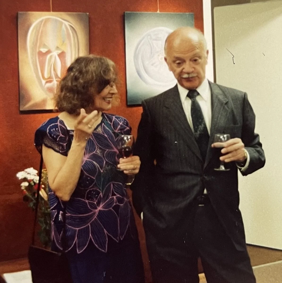 A woman and a man in formal attire each holding a glass of red wine, smiling and conversing at an indoor event. Artworks are hanging on the wall behind them.