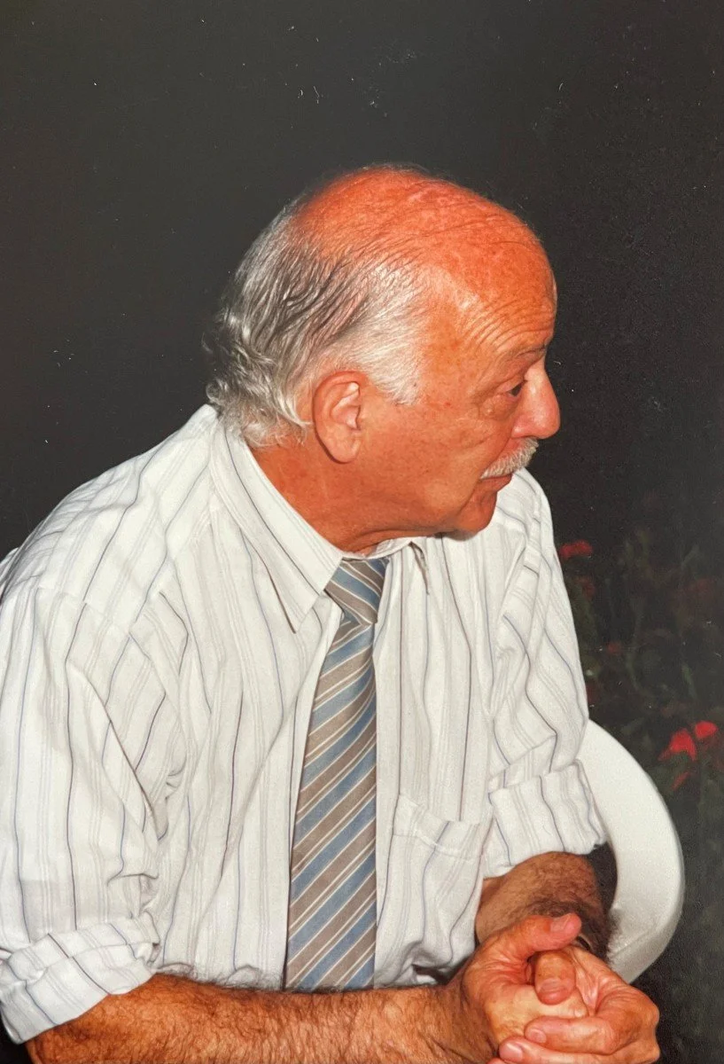 An elderly man with a receding hairline and gray hair, wearing a striped dress shirt and a striped tie, sitting with his hands clasped. The background is dark with some out-of-focus red flowers.