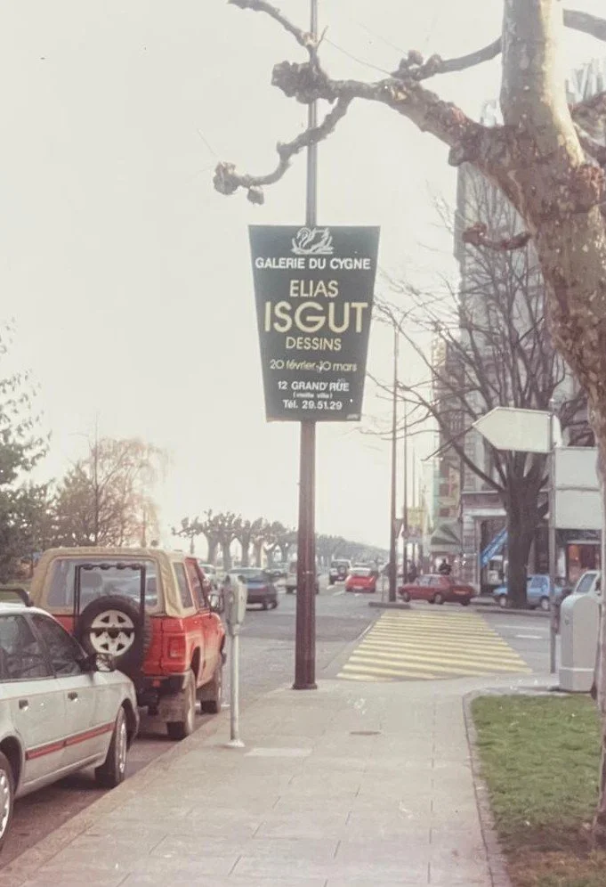 A street view with parked cars and trees on a cloudy day. A sign hanging from a pole advertises an art exhibition at Galerie du Cygne, featuring Elias Isgut, with dates and contact information.