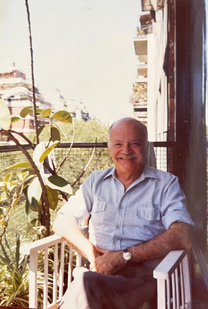 A smiling elderly man with a bald head and beard sitting on a balcony railing, wearing a short-sleeved button-up shirt and a wristwatch. There are green plants nearby, and buildings are visible in the background.