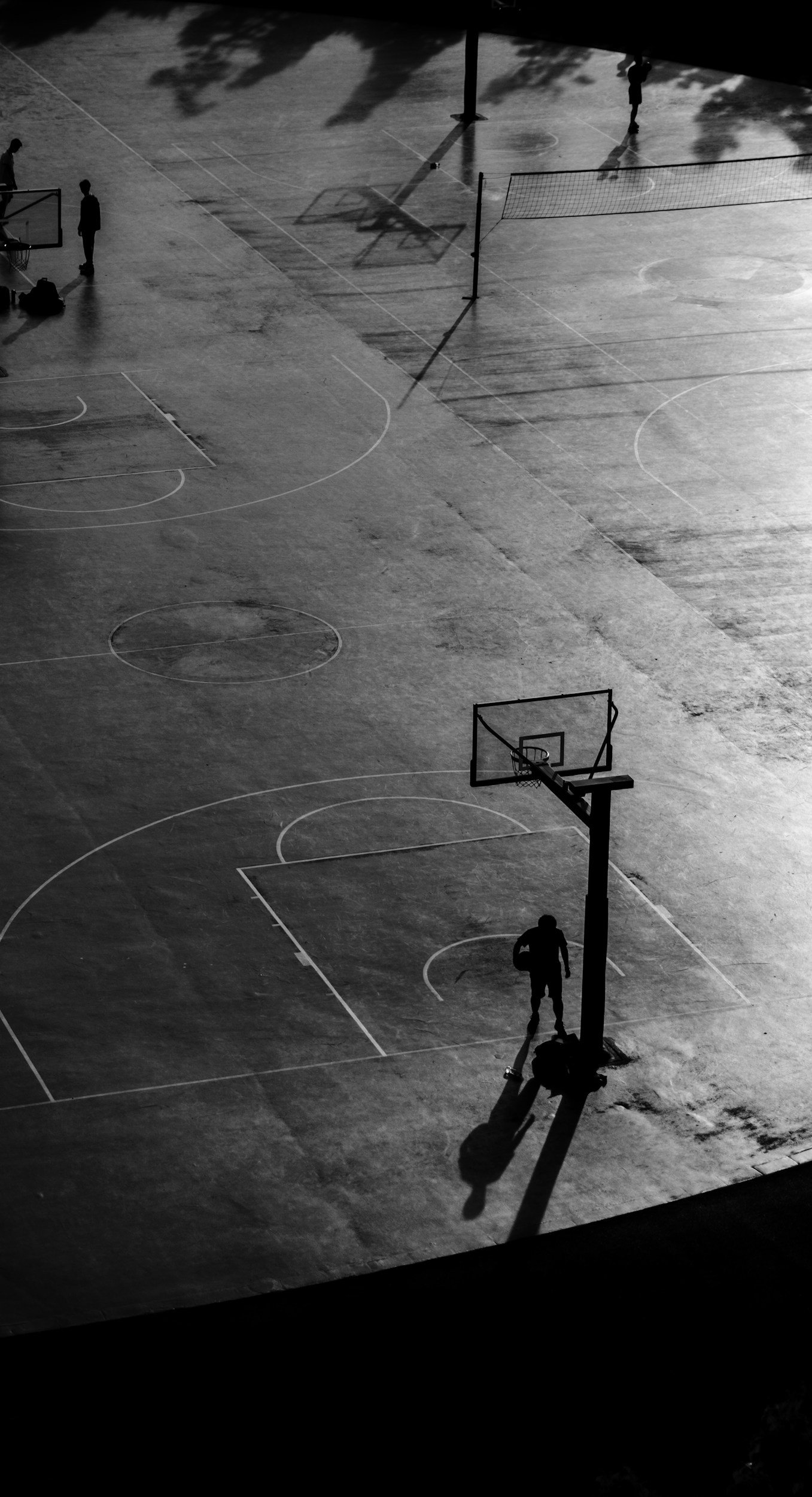 Black and white photo of an outdoor basketball court with a few people playing basketball and shadows of trees cast on the surface.