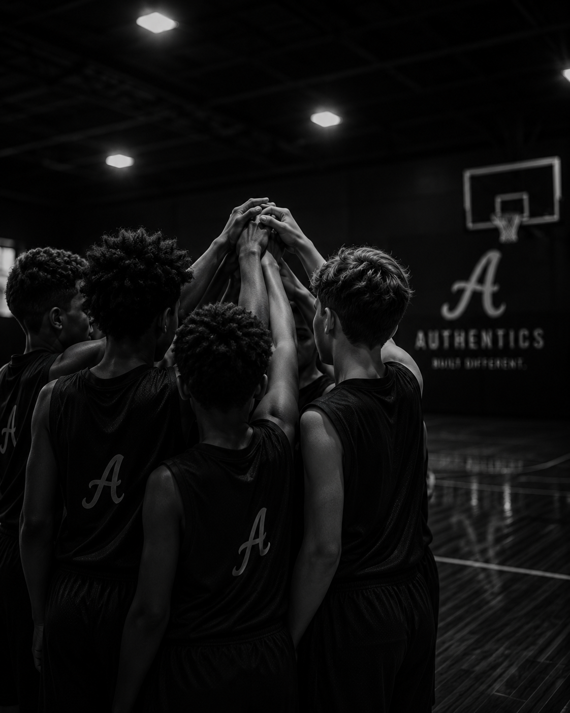 A group of young basketball players in black uniforms forming a huddle with their hands stacked together in an indoor basketball court.