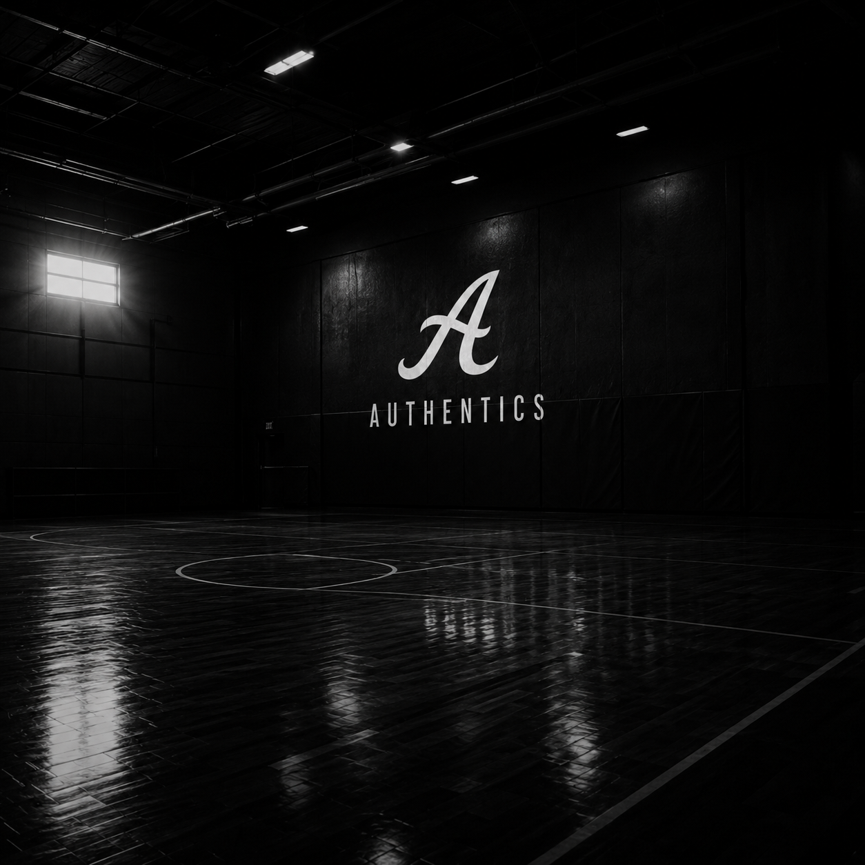Dark indoor basketball court with a large wall that has a white letter "A" and the words "Authentics" written underneath in white, with minimal lighting coming from a window on the left.