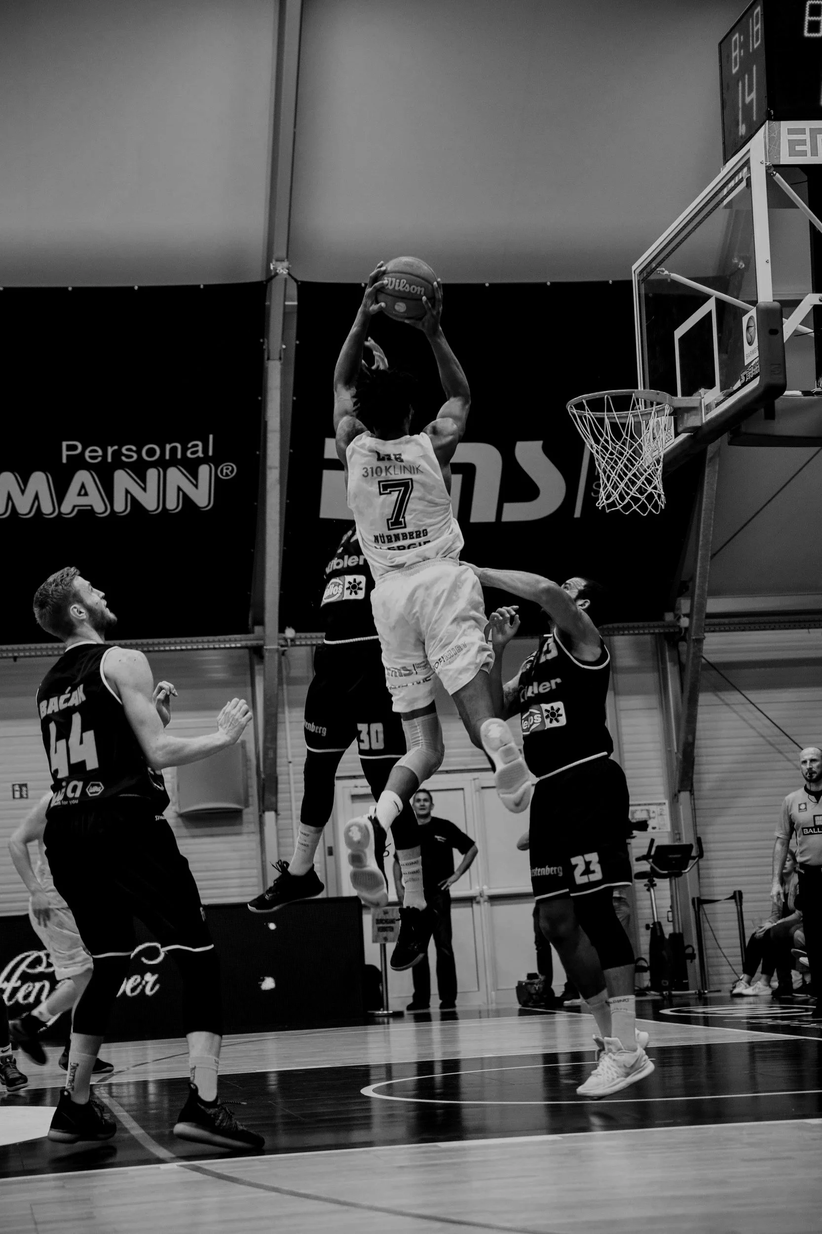 Black and white action shot of a men's basketball game. A player wearing a jersey with the number 7 jumps to make a shot near the basket while others are defending. The gym has banners and digital scoreboards.