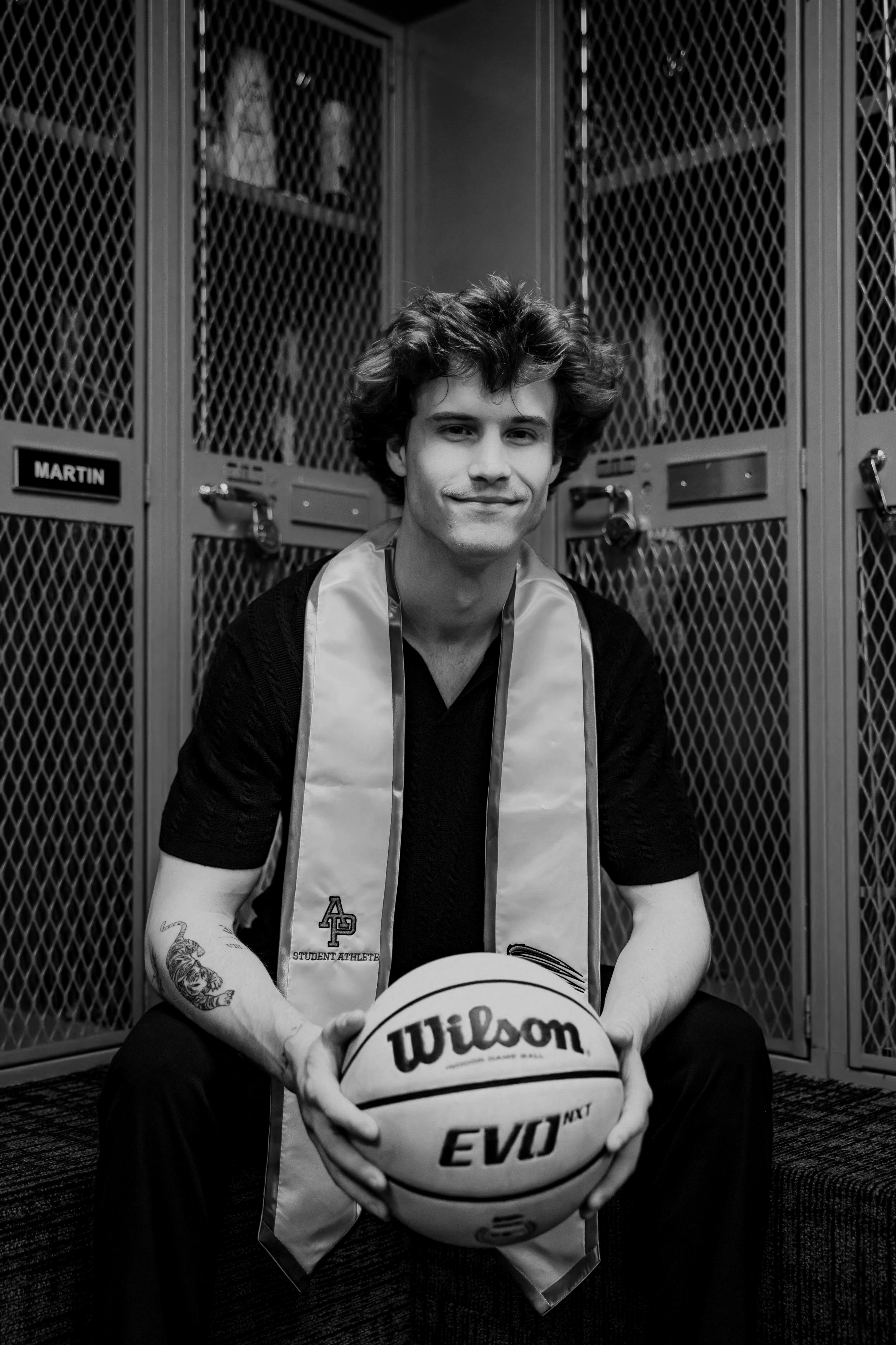 A young man with curly hair and tattoos on his arms, wearing a satin stole and holding a Wilson basketball, sitting in front of lockers in a gym or sports facility.