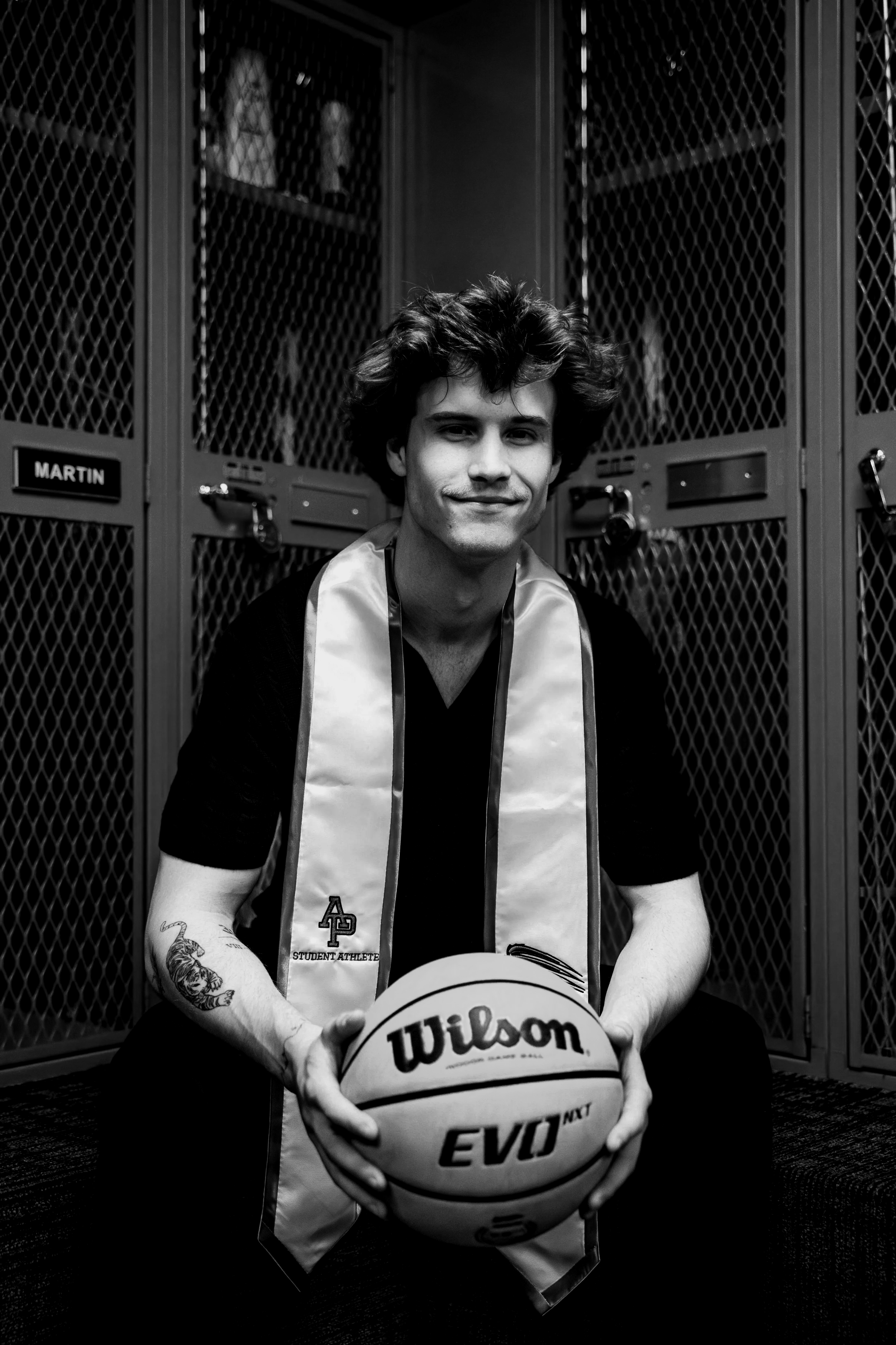 A young man with curly hair and tattoos on his arm, wearing a sports sash, sitting in a locker room, holding a Wilson basketball, and smiling at the camera.