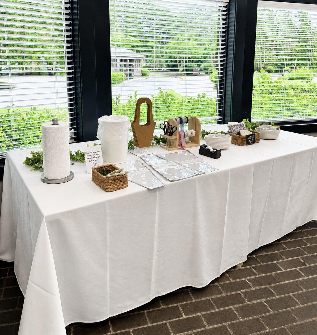 A long table covered with a white cloth, set up near a large window with blinds, displaying various craft supplies including scissors, tape, paper towels, and containers, with greenery visible outside.