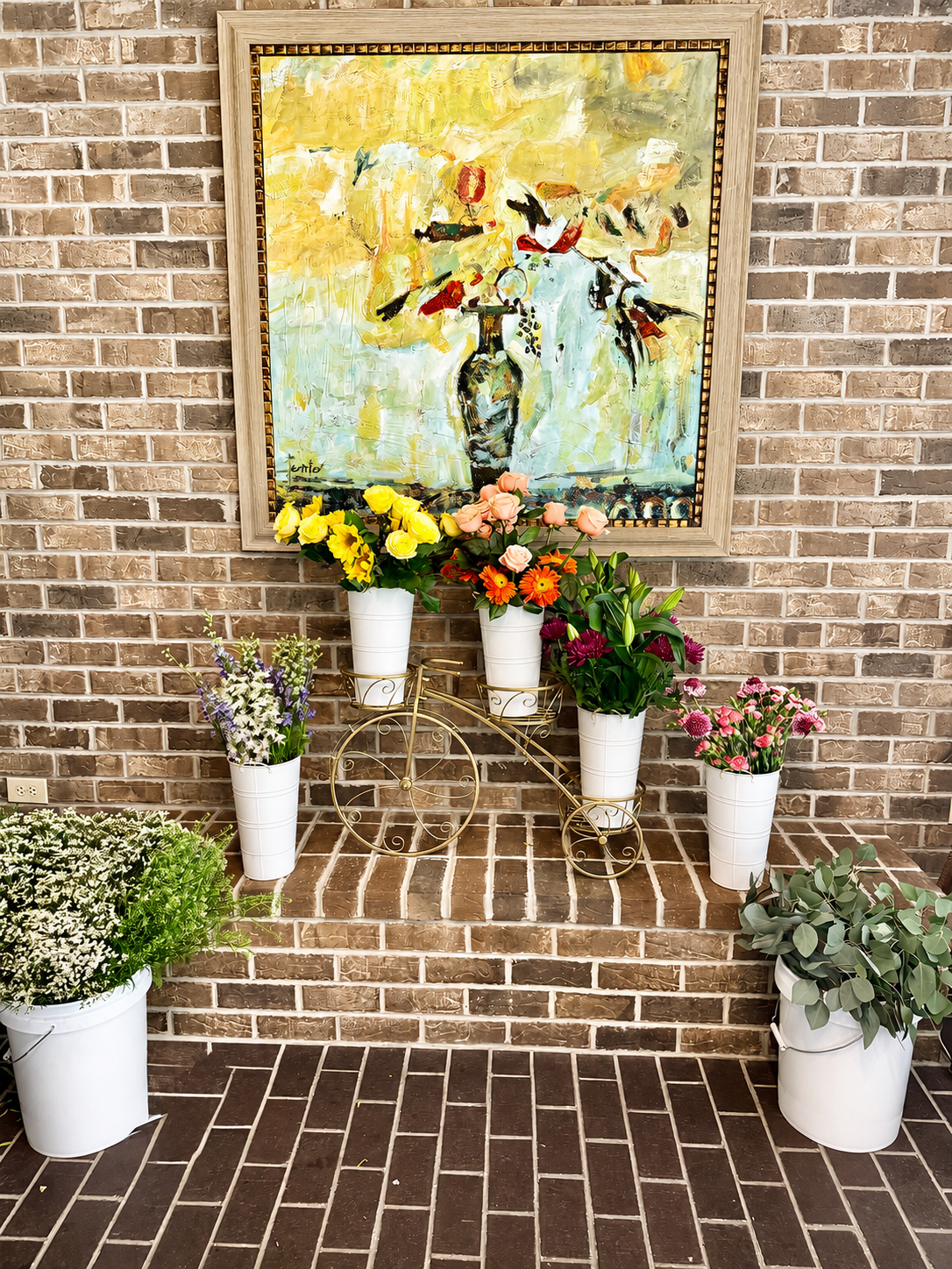 Arrangement of white pots with colorful flowers in front of a brick wall and a framed abstract painting.