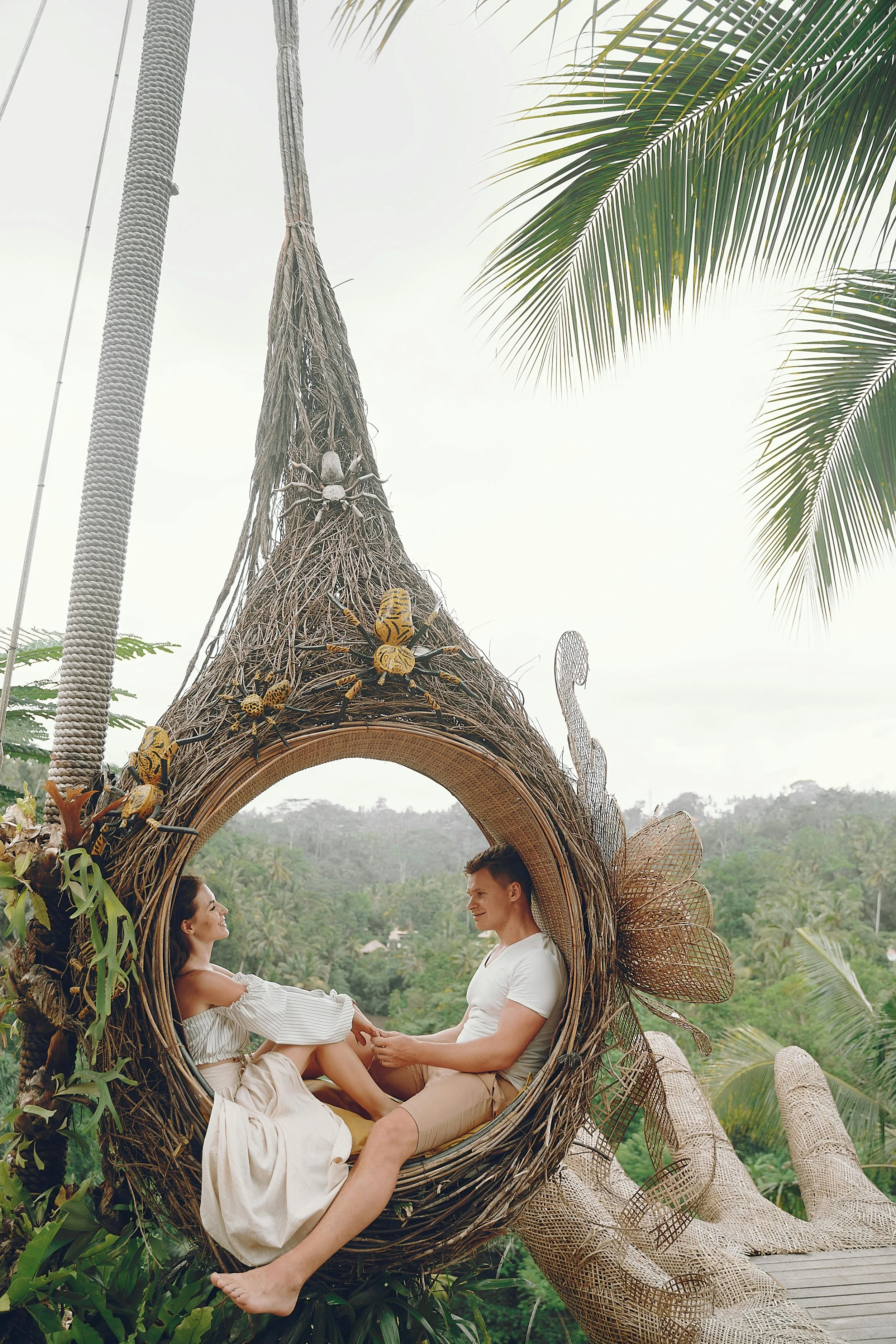 A couple sitting inside a large, nest-like swing made of natural materials, surrounded by lush greenery and palm trees, holding hands and looking at each other.