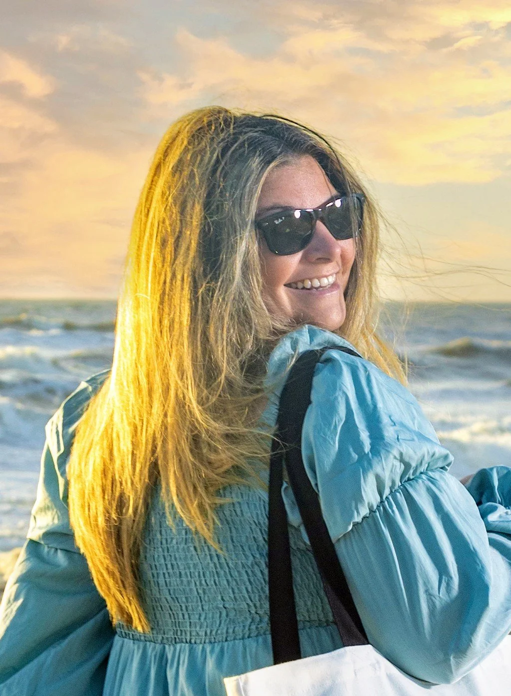 Smiling woman at the beach wearing sunglasses and a blue jacket at sunset.