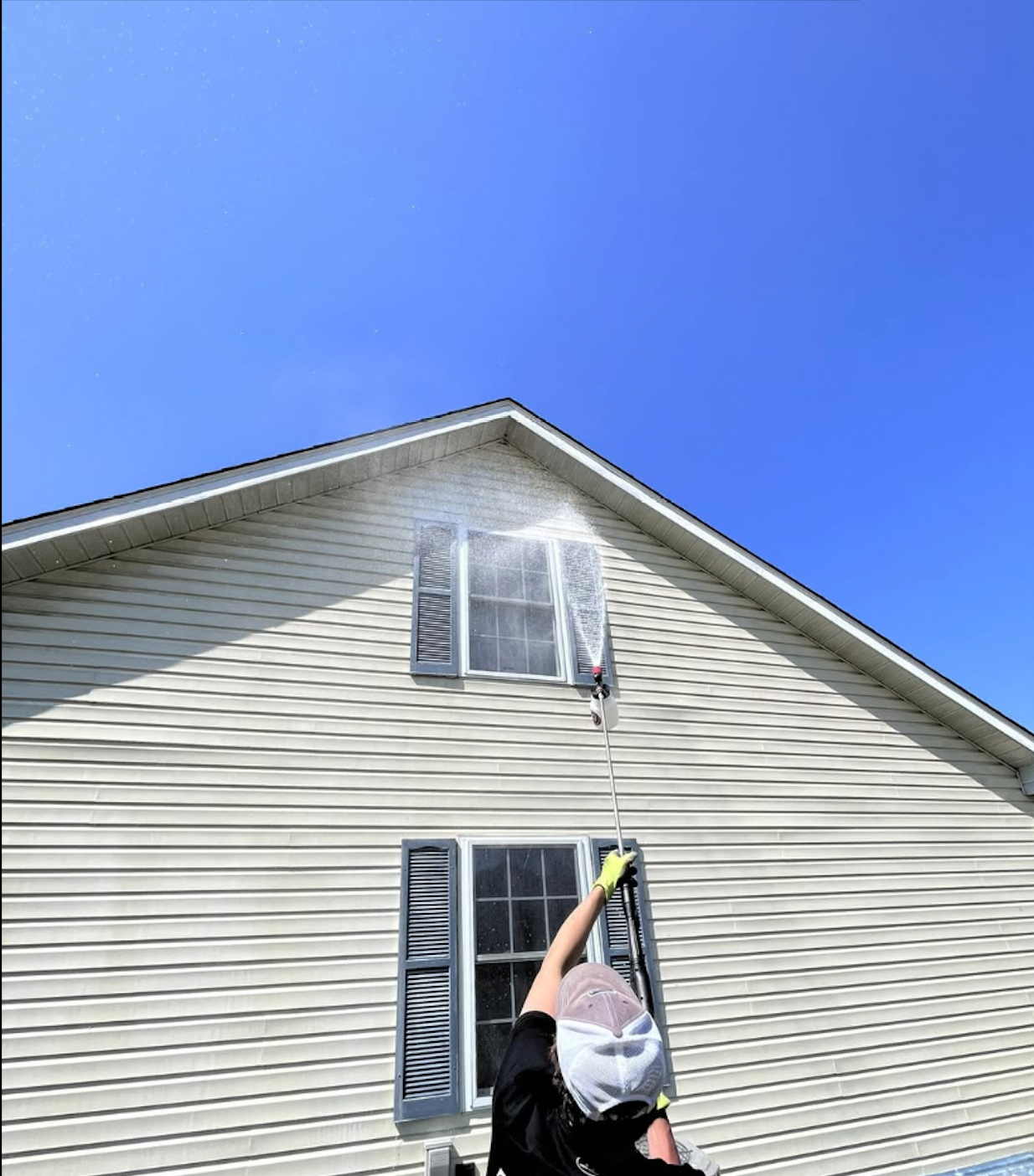 Person cleaning windows on a house with a water spray nozzle, wearing gloves and a hat on a sunny day.
