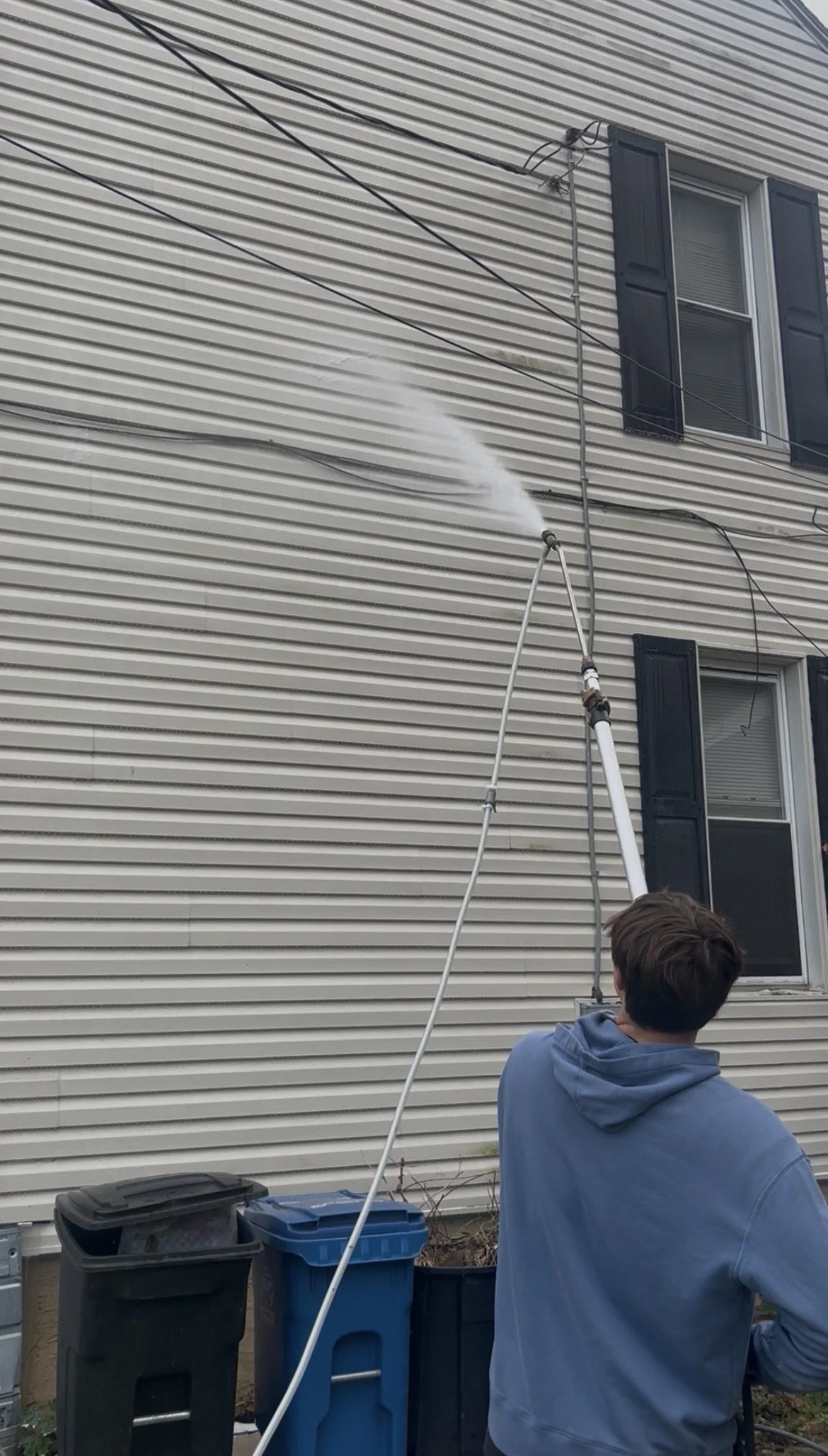 A person in a blue hoodie is pressure washing the beige exterior siding of a house, with water spraying onto the siding. Two trash cans are nearby, and several overhead wires are visible above.