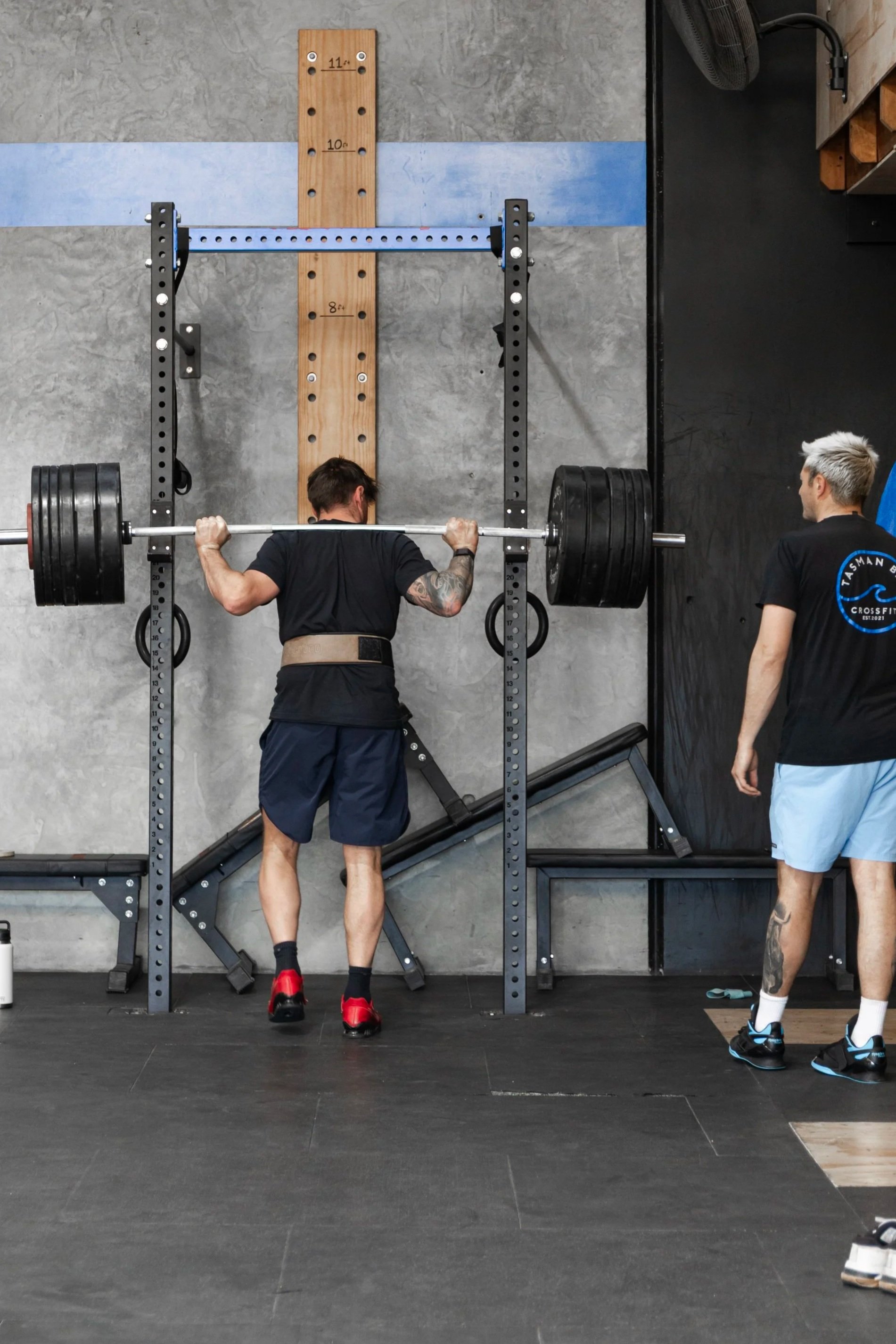 A man performing a barbell squat in a gym, watched by a trainer, with weight plates on the barbell, and a black and gray gym interior with a wall-mounted pegboard and workout equipment.
