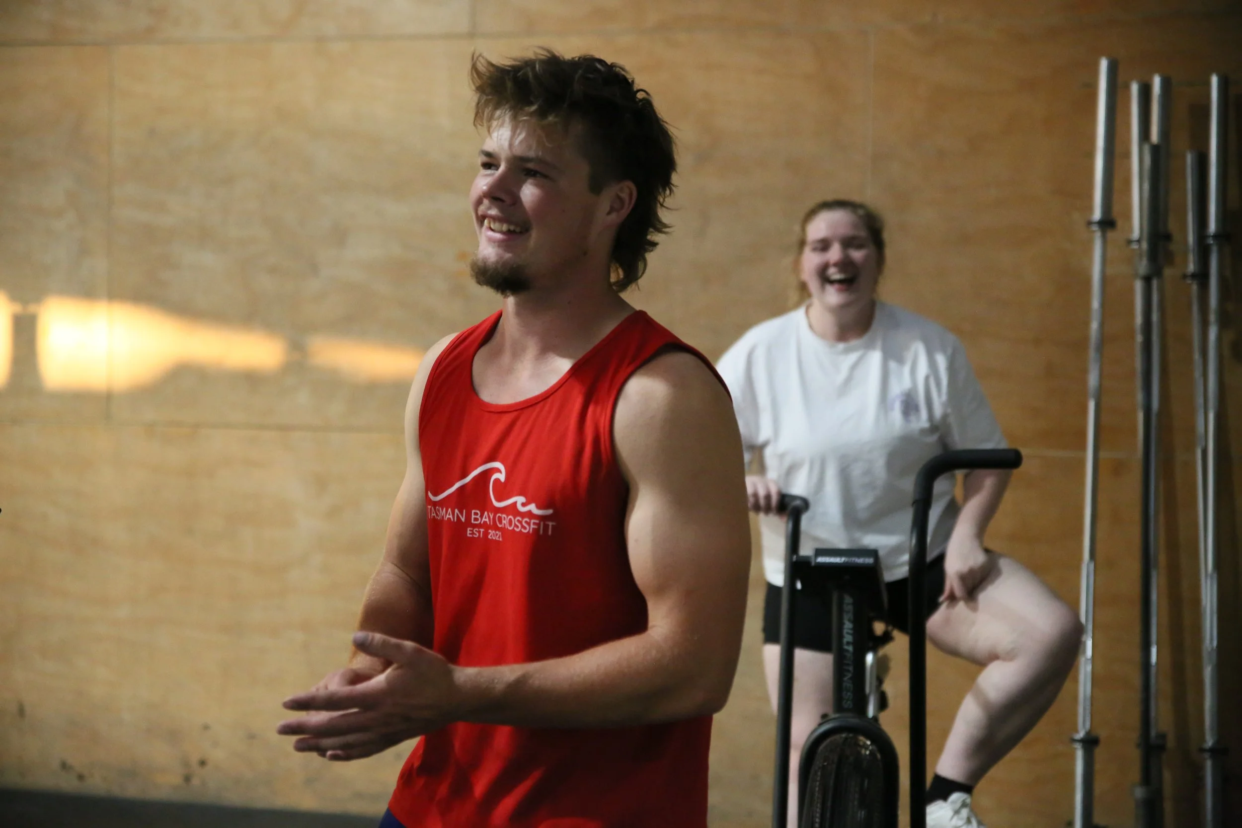A young man in a red tank top smiling and clapping, with a young woman in a wheelchair standing behind him in a gym or fitness facility.