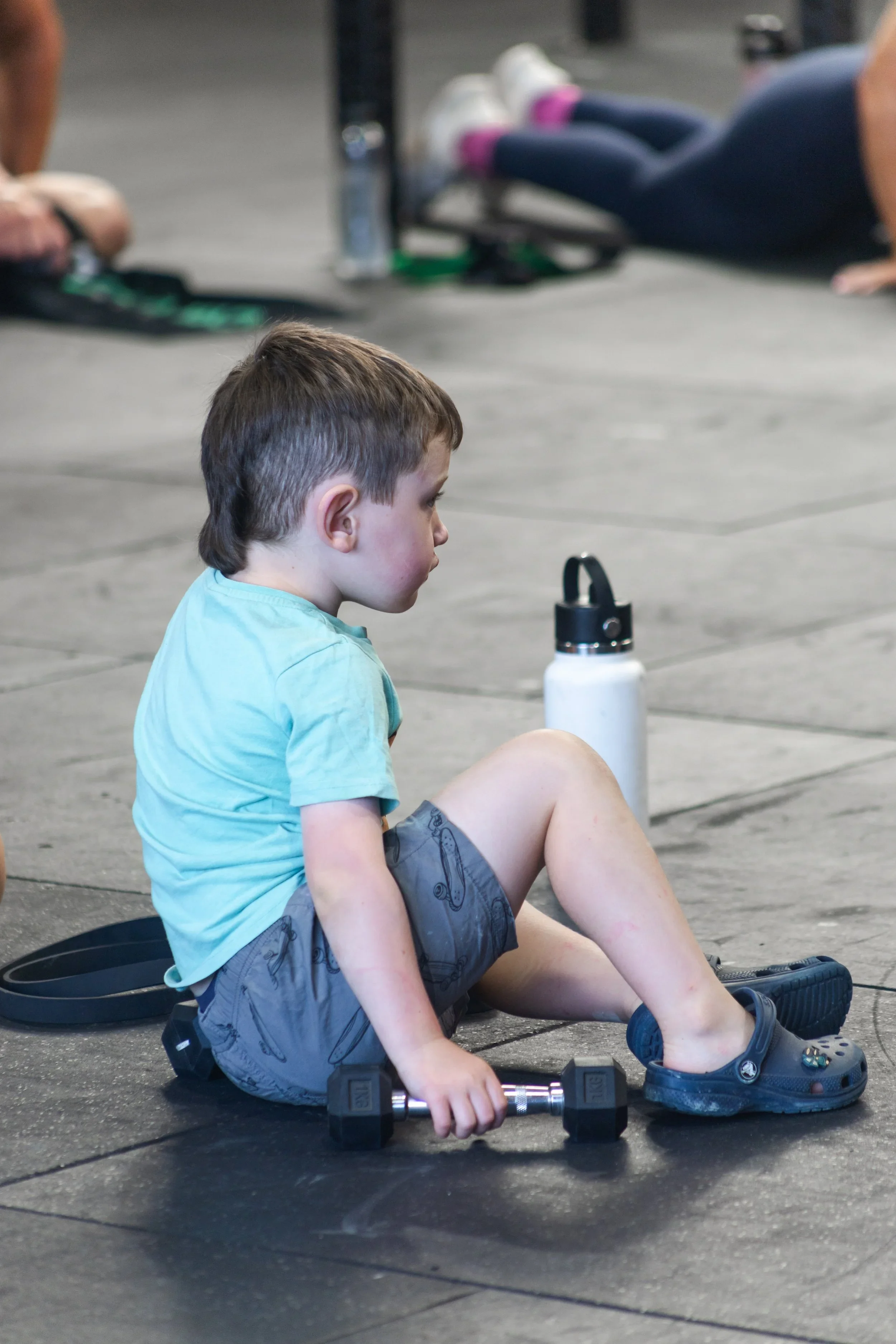 A young boy sitting on the gym floor surrounded by workout equipment, including a water bottle, dumbbells, and a resistance band.
