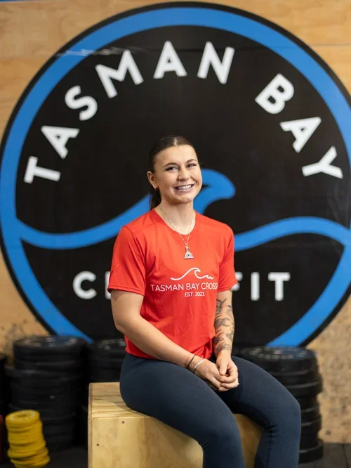 A young woman in a red T-shirt sitting on a wooden block in front of a large circular sign that reads "Tasman Bay" with a stylized wave and bird, inside a gym or crossfit box.