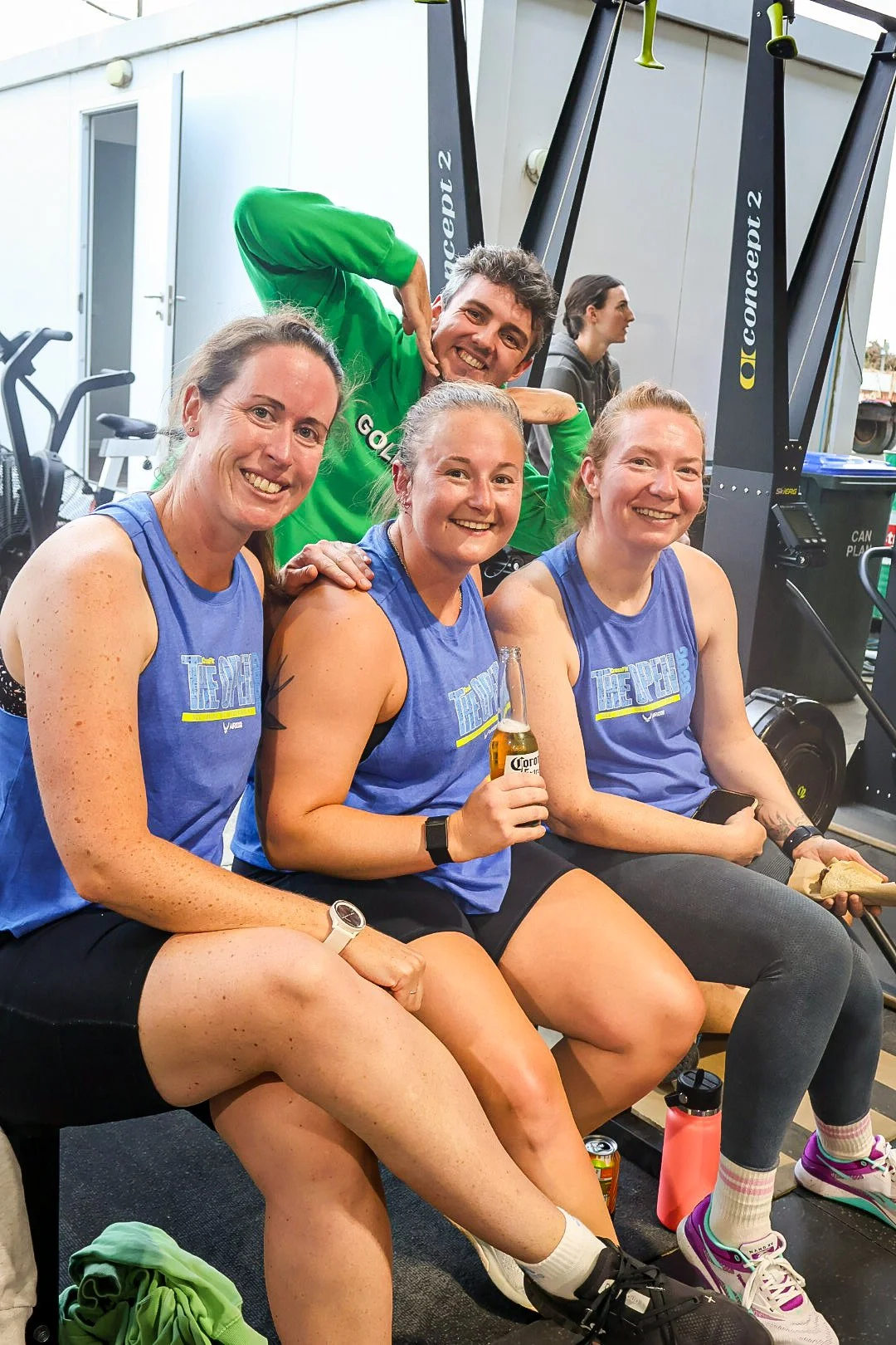 Four women in blue athletic shirts and a man in a green jacket smiling and sitting on a bench after a workout, with gym equipment in the background.