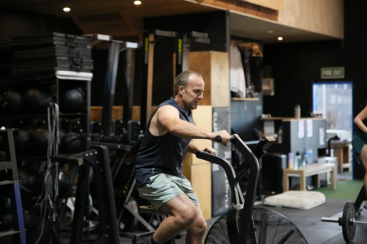 A man exercising on a fan bike in a gym, surrounded by workout equipment and weights.