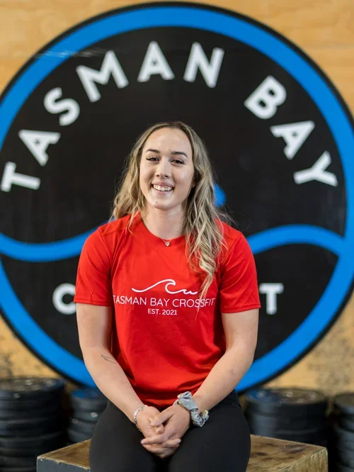 A woman in a red t-shirt sitting in front of a Tasman Bay CrossFit sign, smiling at the camera.
