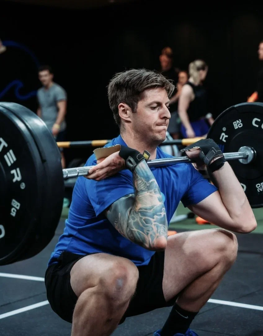 Man squatting and lifting a barbell in a gym with other people in the background.