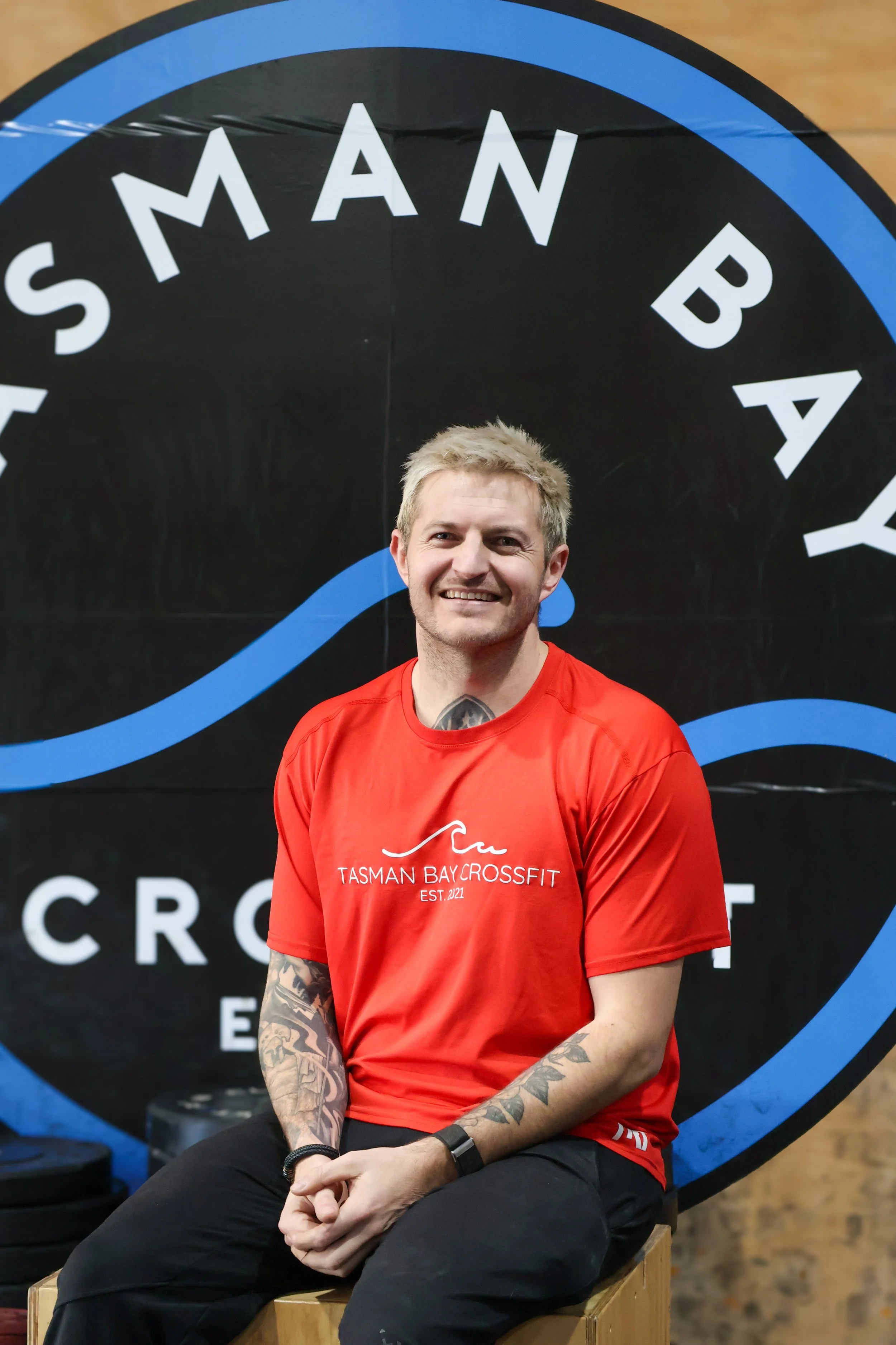 A smiling man with short blonde hair and tattoos on his left arm, wearing a red Tasma Bay Crossfit shirt, sitting on a wooden box in front of a black and blue CrossFit logo background.