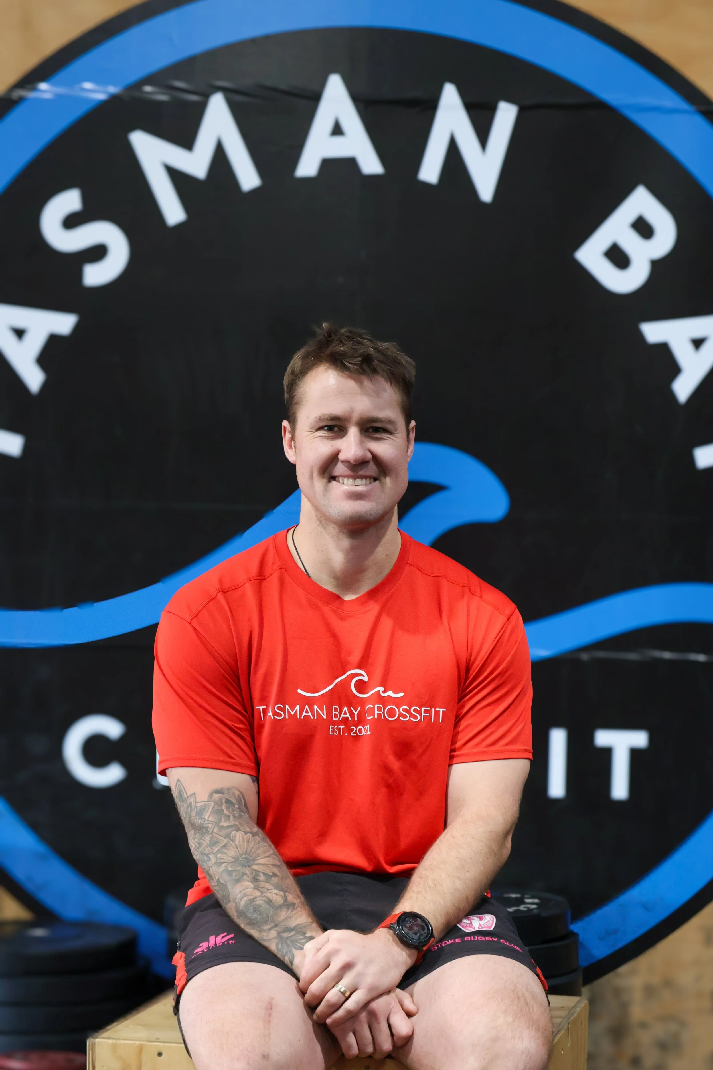 A smiling young man sitting with crossed arms, wearing a red T-shirt with 'Tasman Bay Crossfit' printed on it, sitting on a wooden box in a gym with a black and blue logo backdrop.
