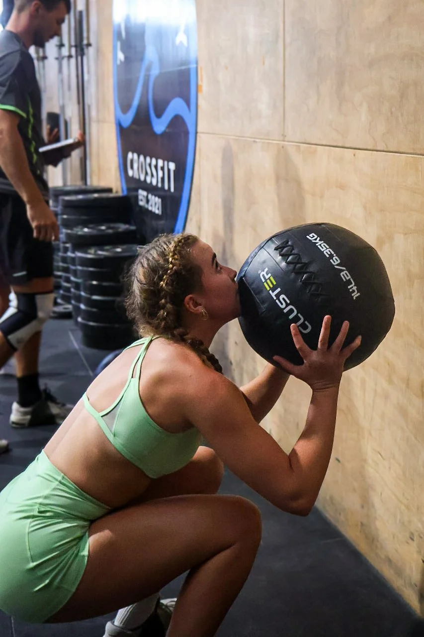 A woman in a green sports bra and shorts squatting and holding a medicine ball in front of her mouth during a workout at a gym. Behind her, a man is standing near a stack of weight plates, and a large CrossFit banner is visible on the wall.