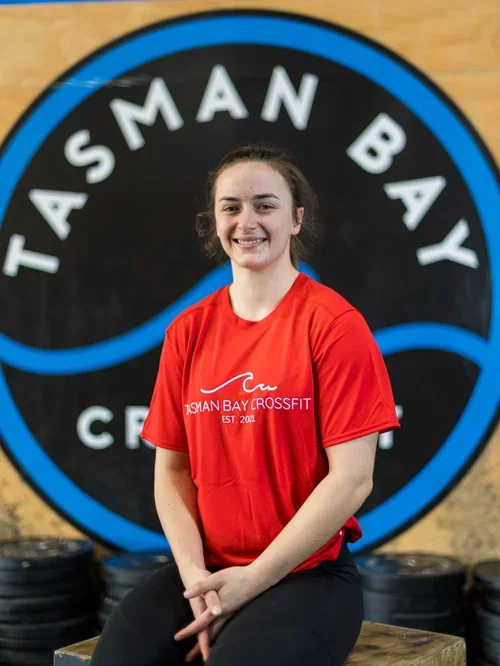 A young woman smiling at the camera, wearing a red Tasman Bay Crossfit t-shirt, seated in front of a large Tasman Bay Crossfit logo on the wall.