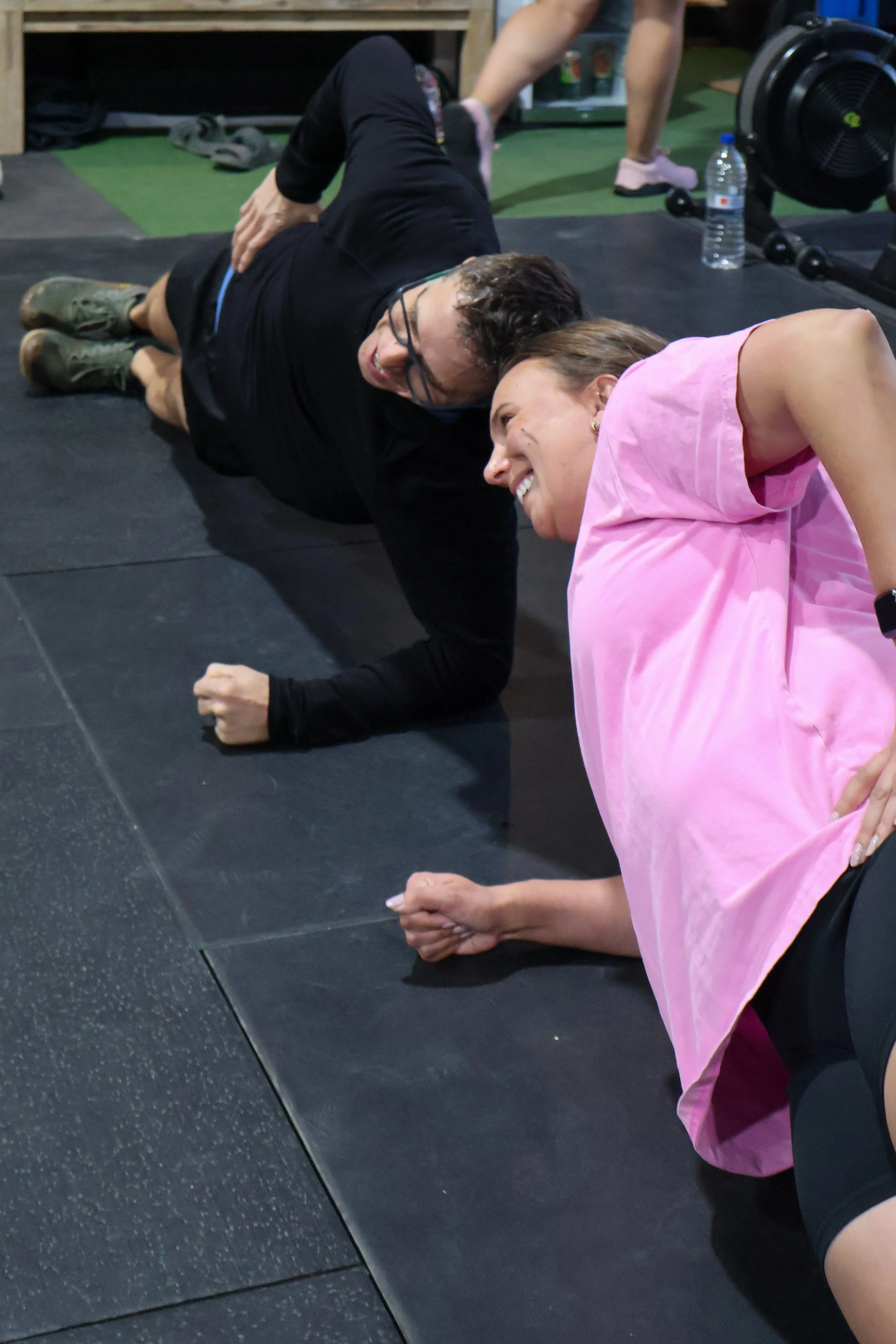 Two women are working out on the floor, doing side planks. One woman is wearing a pink top and the other is in black.