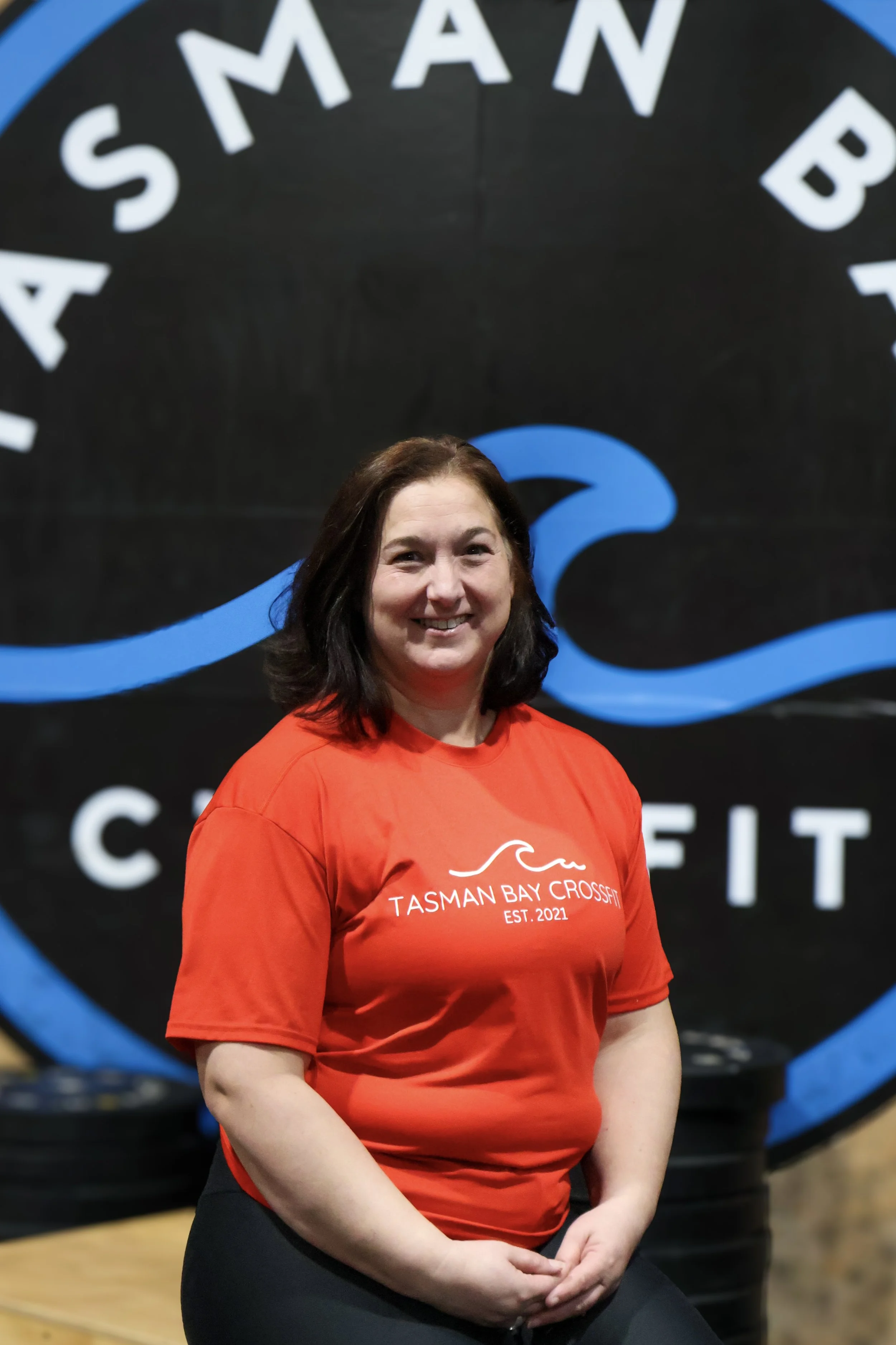 A smiling woman with dark hair and light skin, wearing a red T-shirt with white text, standing in front of a large black and blue logo that says 'Tasman Bay Crossfit.' She is indoors, with her hands resting on her lap.