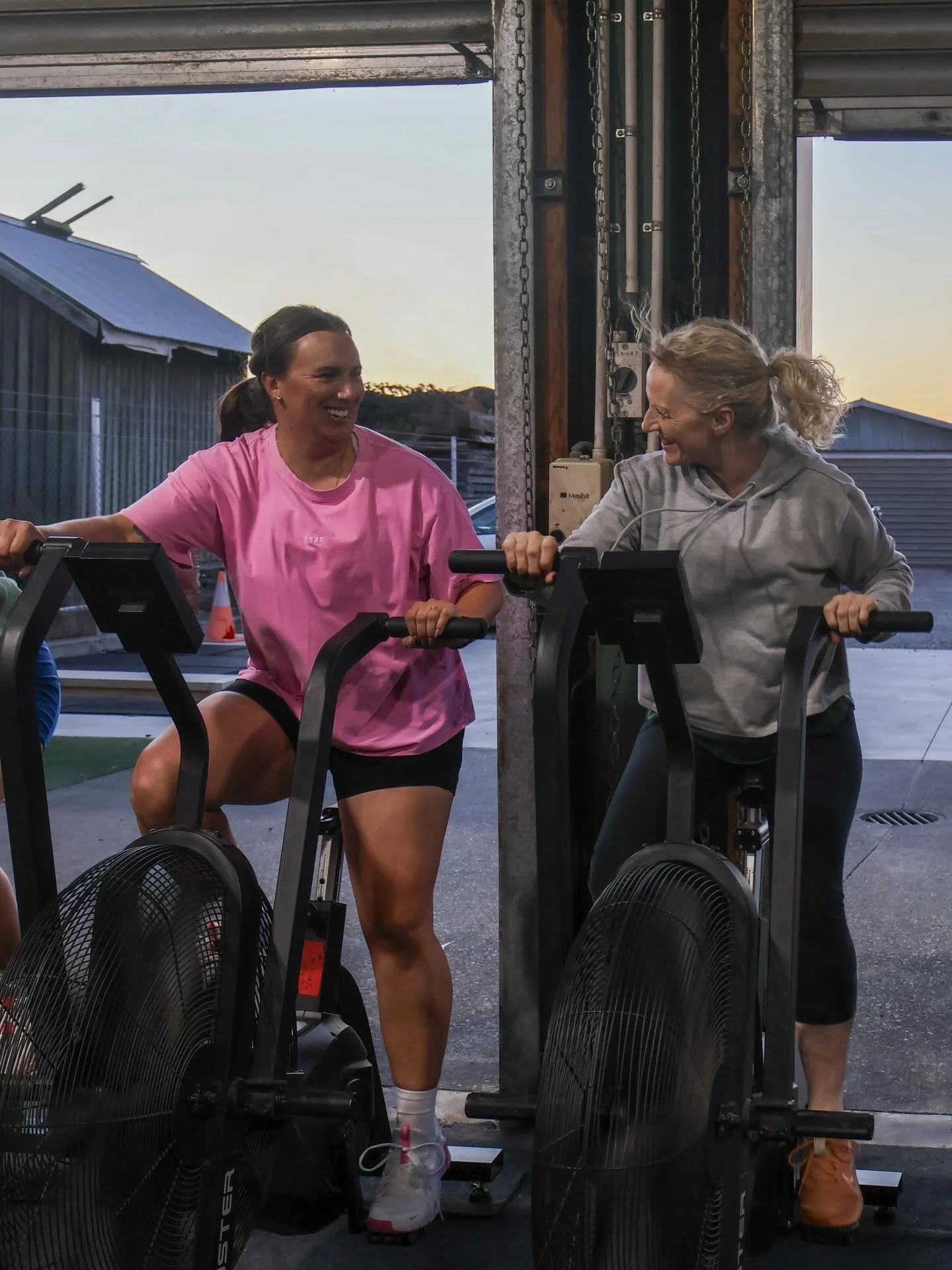 Two women working out on assault bikes in a gym, smiling and facing each other, with sunset view outside.