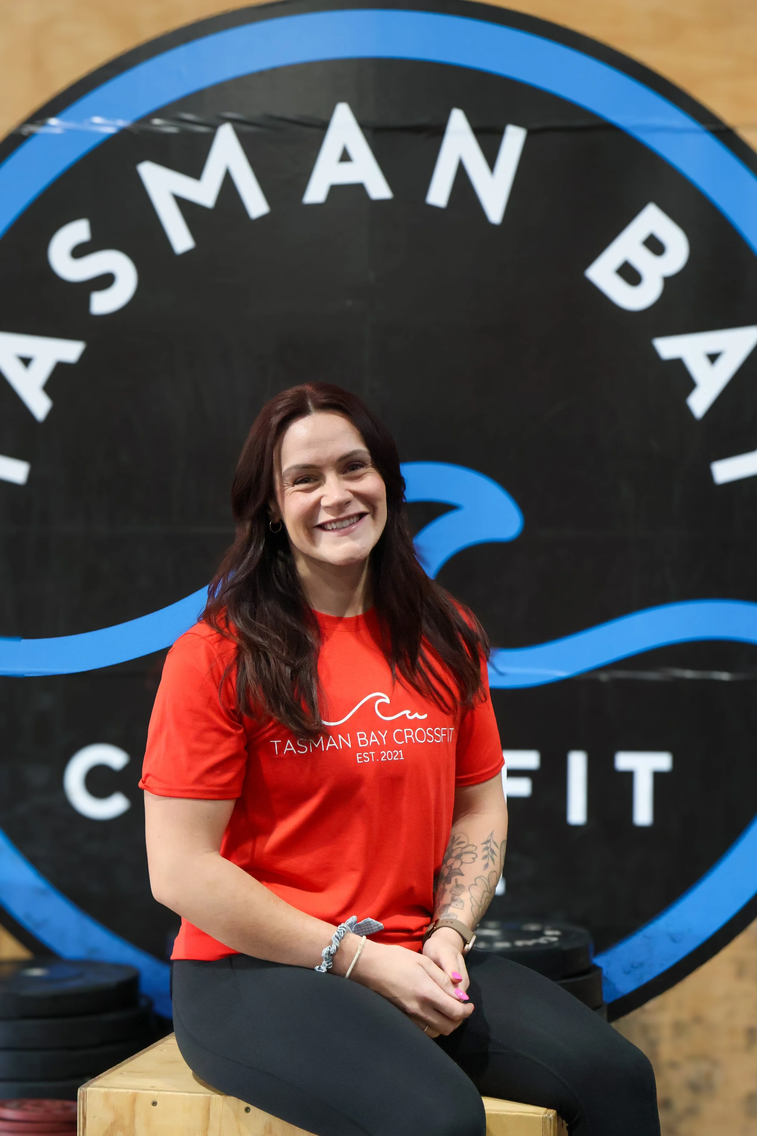 A woman with long dark hair wearing a red Tasman Bay Crossfit t-shirt, sitting on a wooden box in a gym, smiling at the camera, with a black and blue logo in the background.