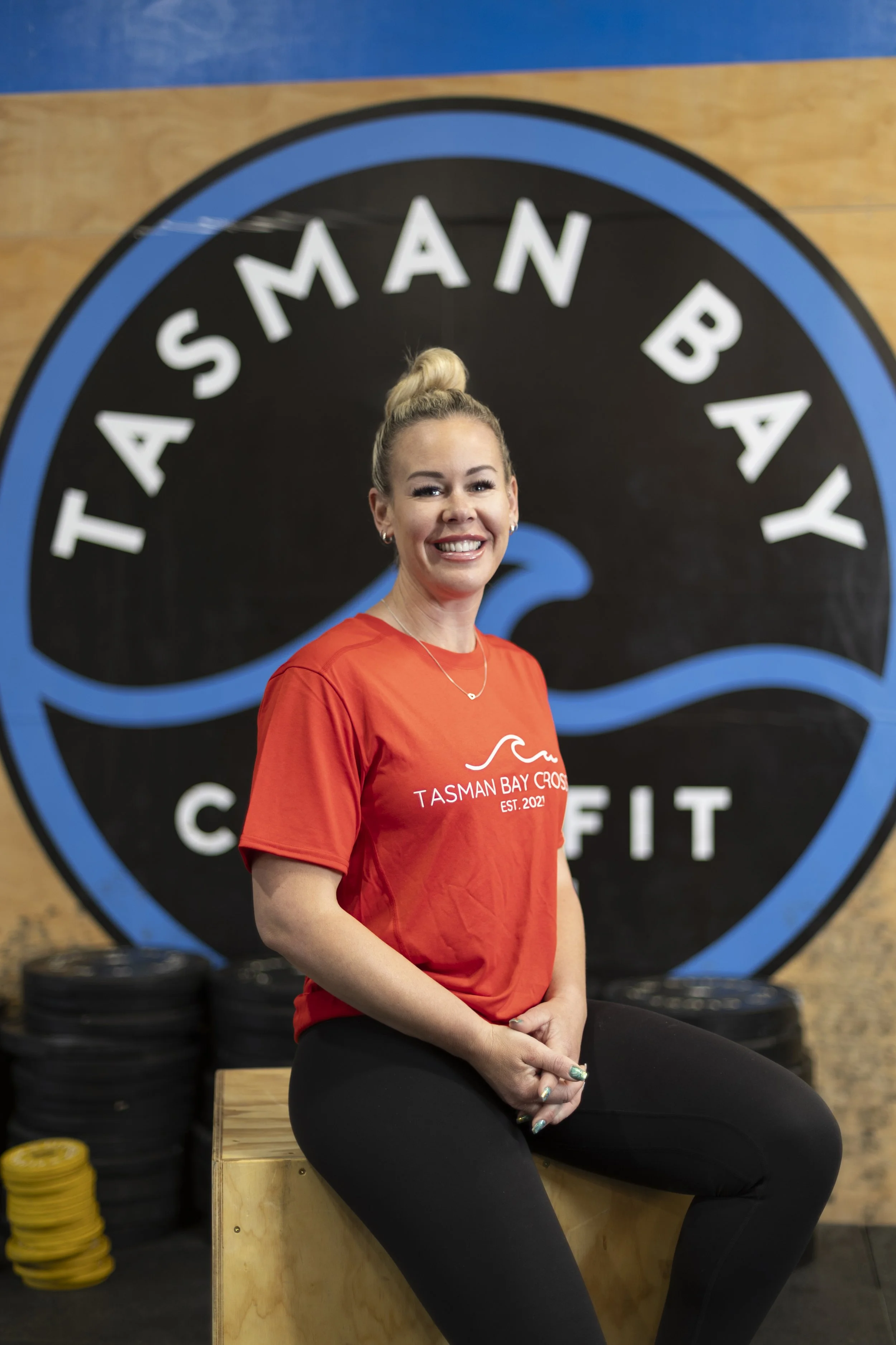 A woman with blonde hair tied in a bun, wearing a red Tasman Bay Crossfit T-shirt, sitting on a wooden box inside a gym, smiling at the camera.