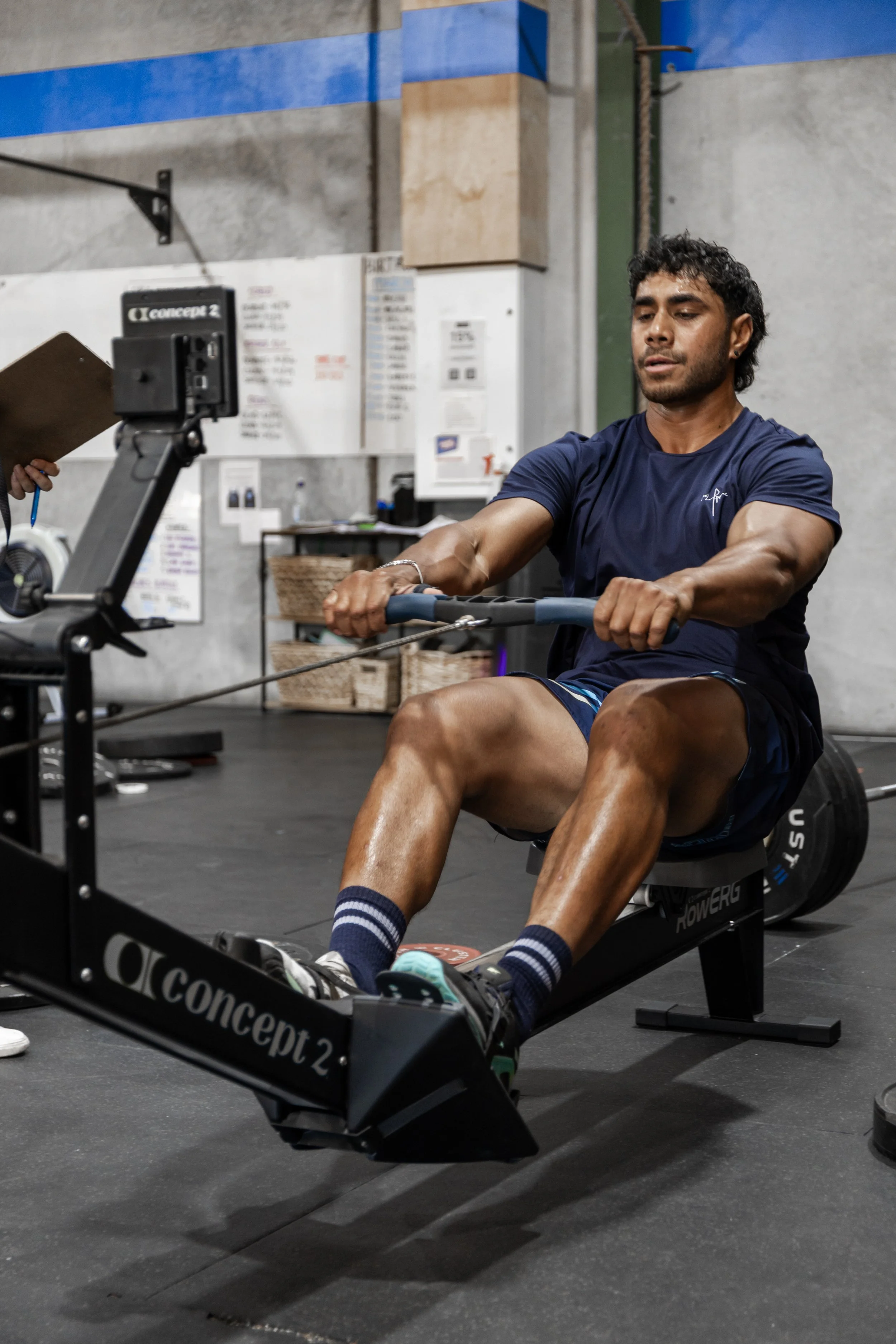 A man with dark curly hair and a beard is seated on a rowing machine, exercising in a gym.