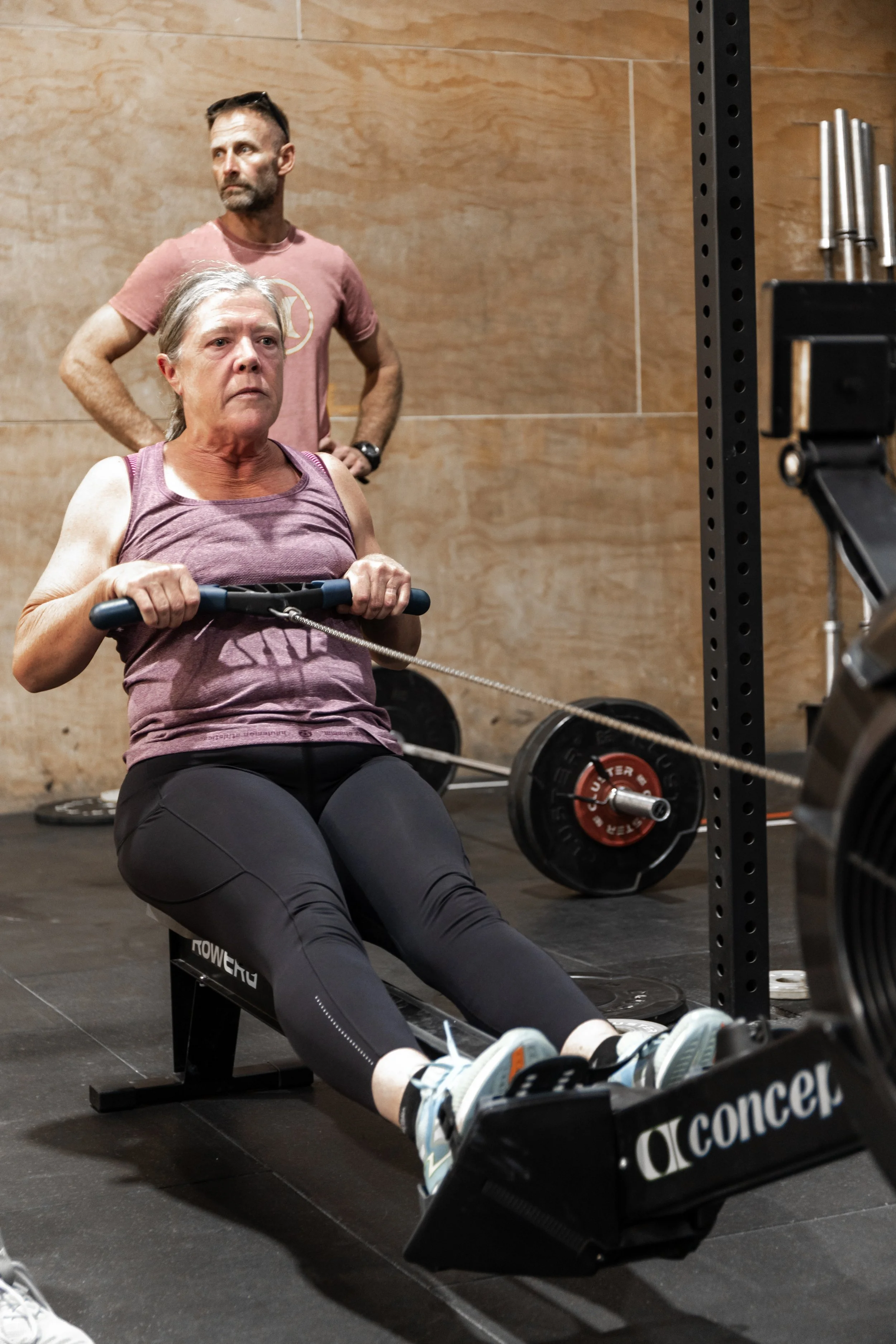 An older woman in workout clothes using a rowing machine in a gym, with a man standing behind her observing.