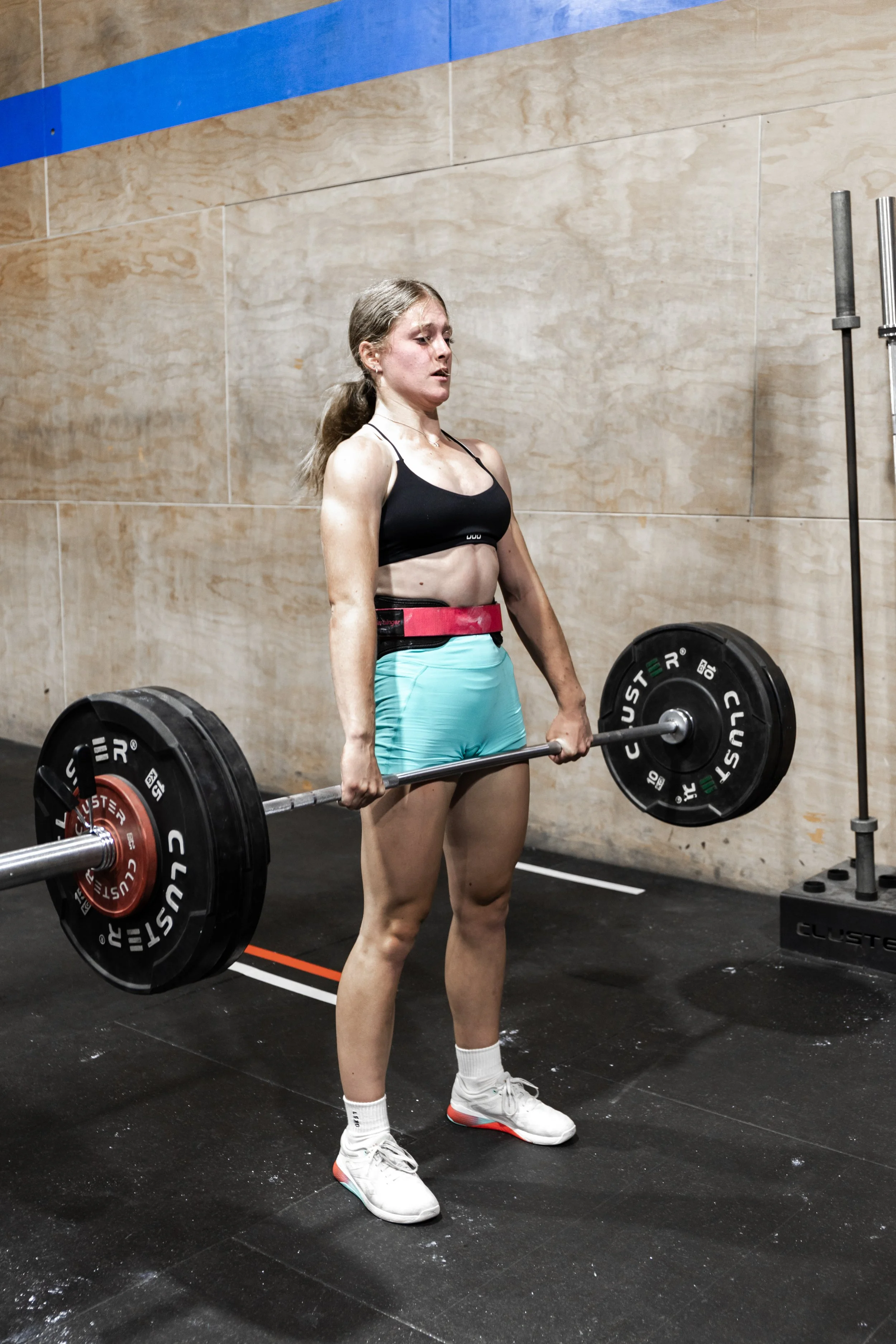 A woman in athletic clothing lifting a barbell with weights in a gym. She is wearing a black sports bra, teal shorts, white sneakers, and white socks, standing on a black gym floor.