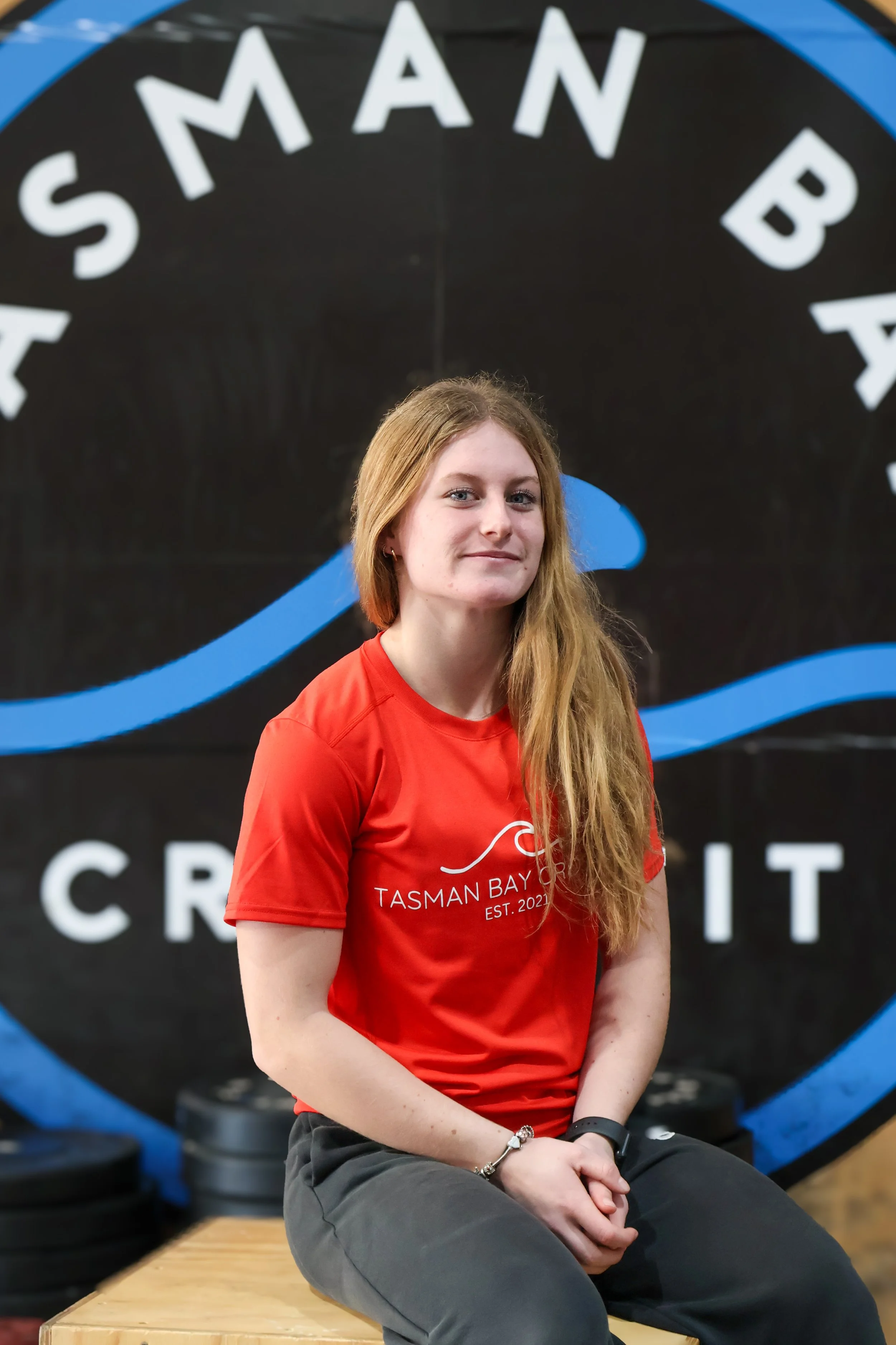 A young woman with long red hair sitting on a wooden bench in front of a black and blue wall with the words Tasman Bay Crossfit behind.