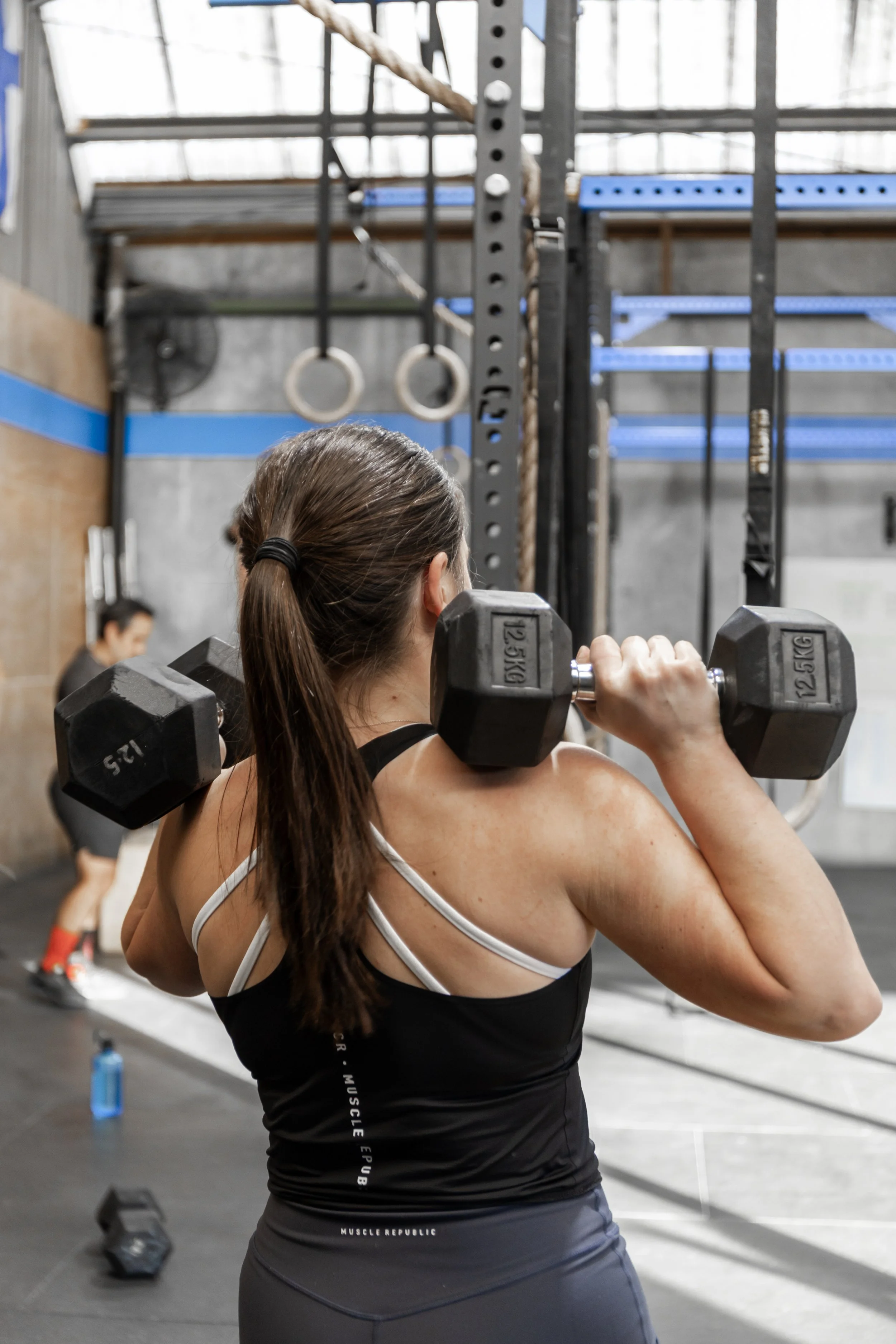 A woman lifting dumbbells in a gym, with gym equipment and another person in the background.