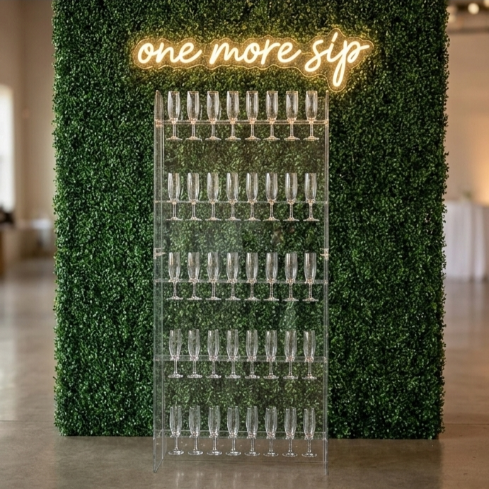 A clear acrylic stand holding multiple empty champagne flutes against a lush green hedge backdrop with a neon sign that reads 'one more sip'.