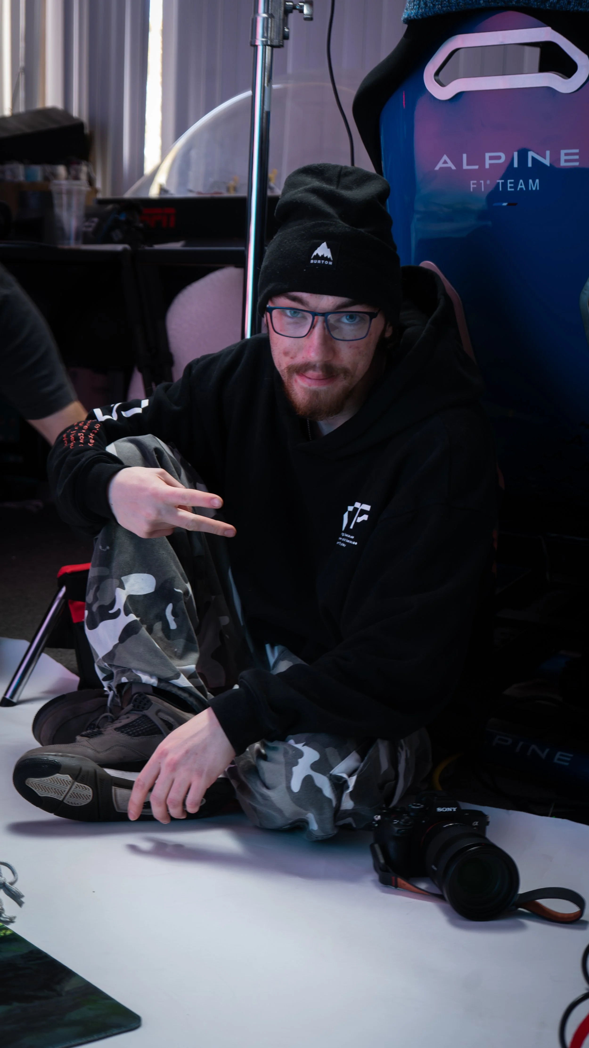 Young man wearing glasses, a black beanie with a mountain logo, and a black hoodie sitting at a table with a camera in front of him, making a peace sign with his right hand, in a room with a blue racing seat and a Alpine F1 Team poster in the background.