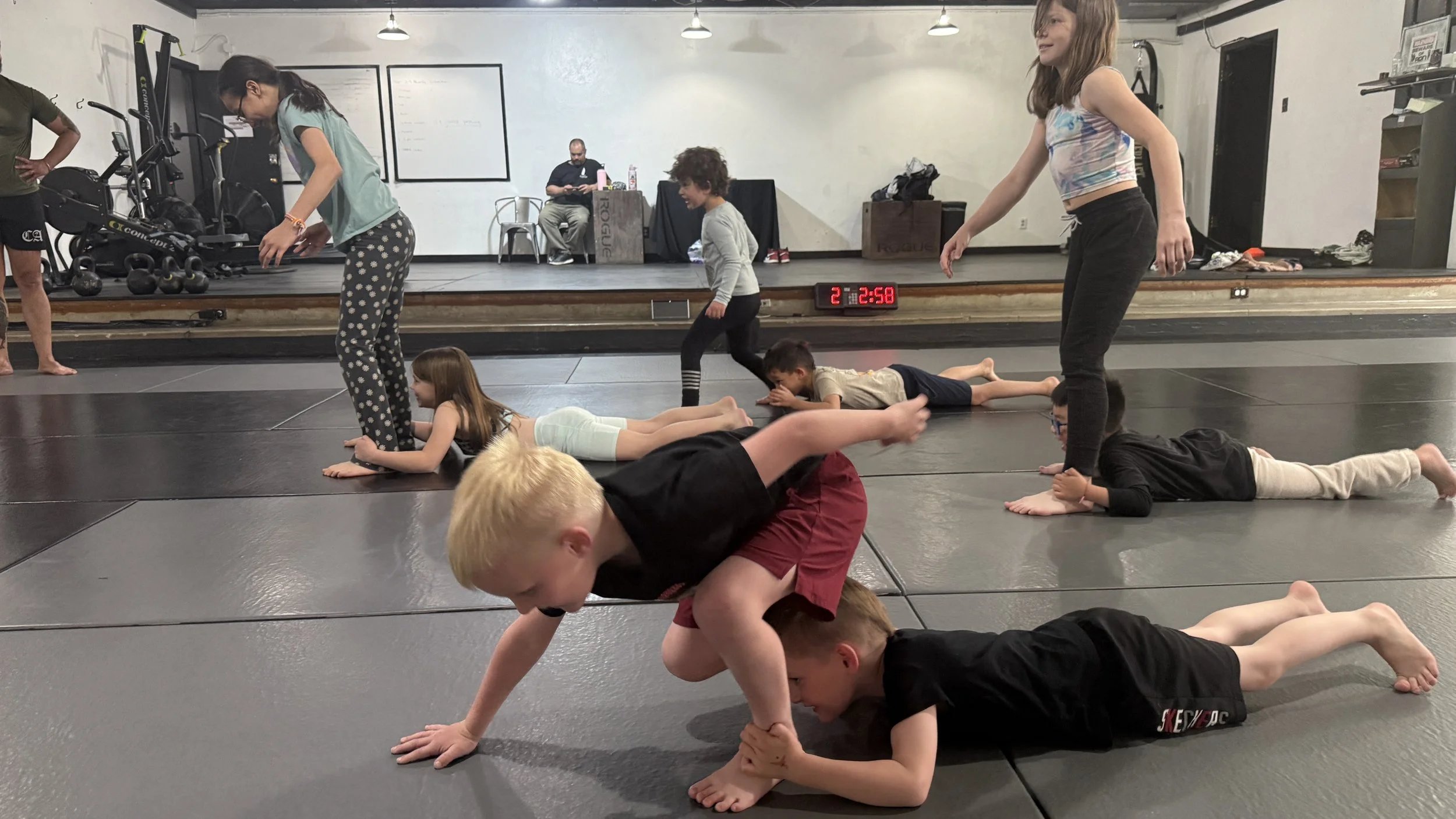 Children practicing a plank exercise on martial arts mats in a gym, supervised by instructors, with a digital timer on the wall showing 2 minutes and 58 seconds.