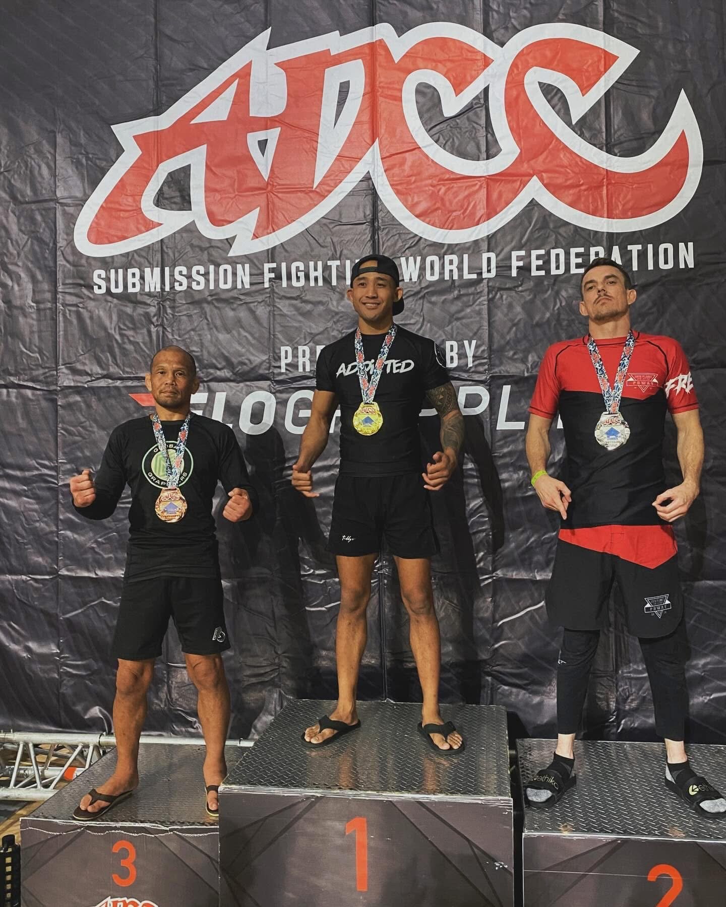 Three men standing on a winners' podium with medals around their necks, in front of a black backdrop with the words 'ADCC Submission Fighting World Federation'. The central man is in first place on the highest platform, wearing a black T-shirt and shorts, smiling. The man on the left is in third place, wearing a black compression shirt and shorts, making a fist. The man on the right is in second place, wearing a red and black shirt and black pants with fists clenched.