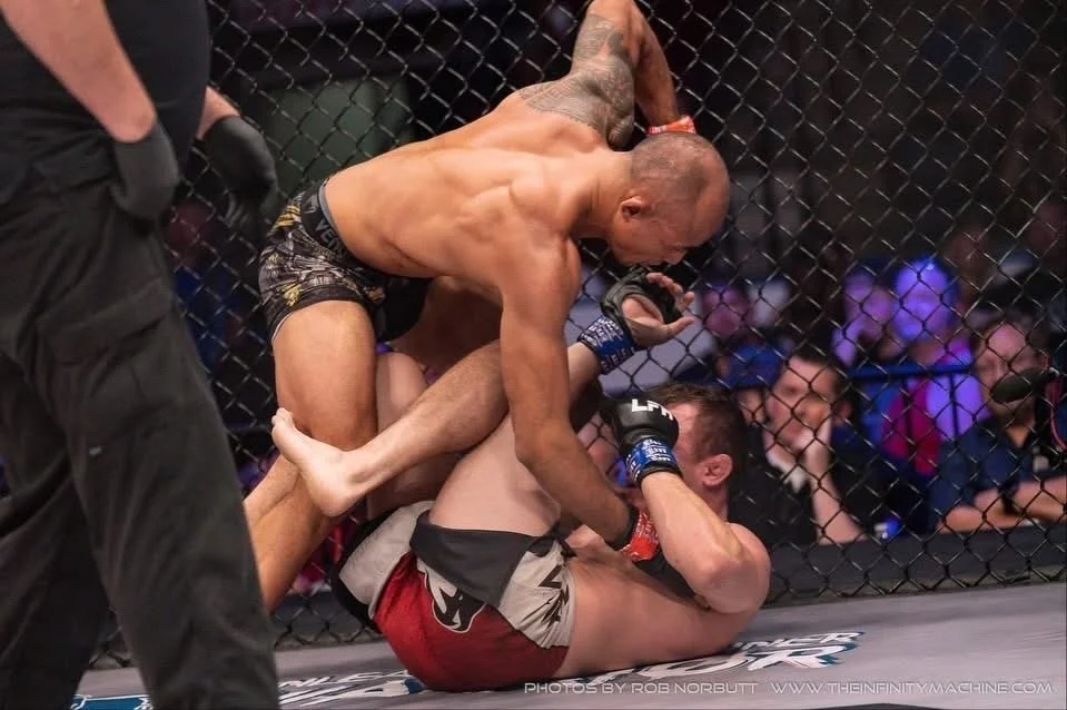 Two mixed martial artists inside a cage, one on top of the other, engaged in a ground fight. The fighter on top is wearing black and gold shorts and has tattoos, while the fighter on the bottom is green shorts with red and black. A referee is partially visible on the left side of the image, observing the fight.