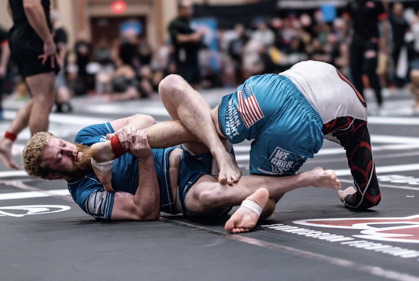 Two male wrestlers competing on a black mat in a gymnasium. One wrestler with a beard and curly hair is on the ground, while the other in a white shirt and blue shorts is on top, grabbing his opponent. Blurred spectators and other competitors are in the background.