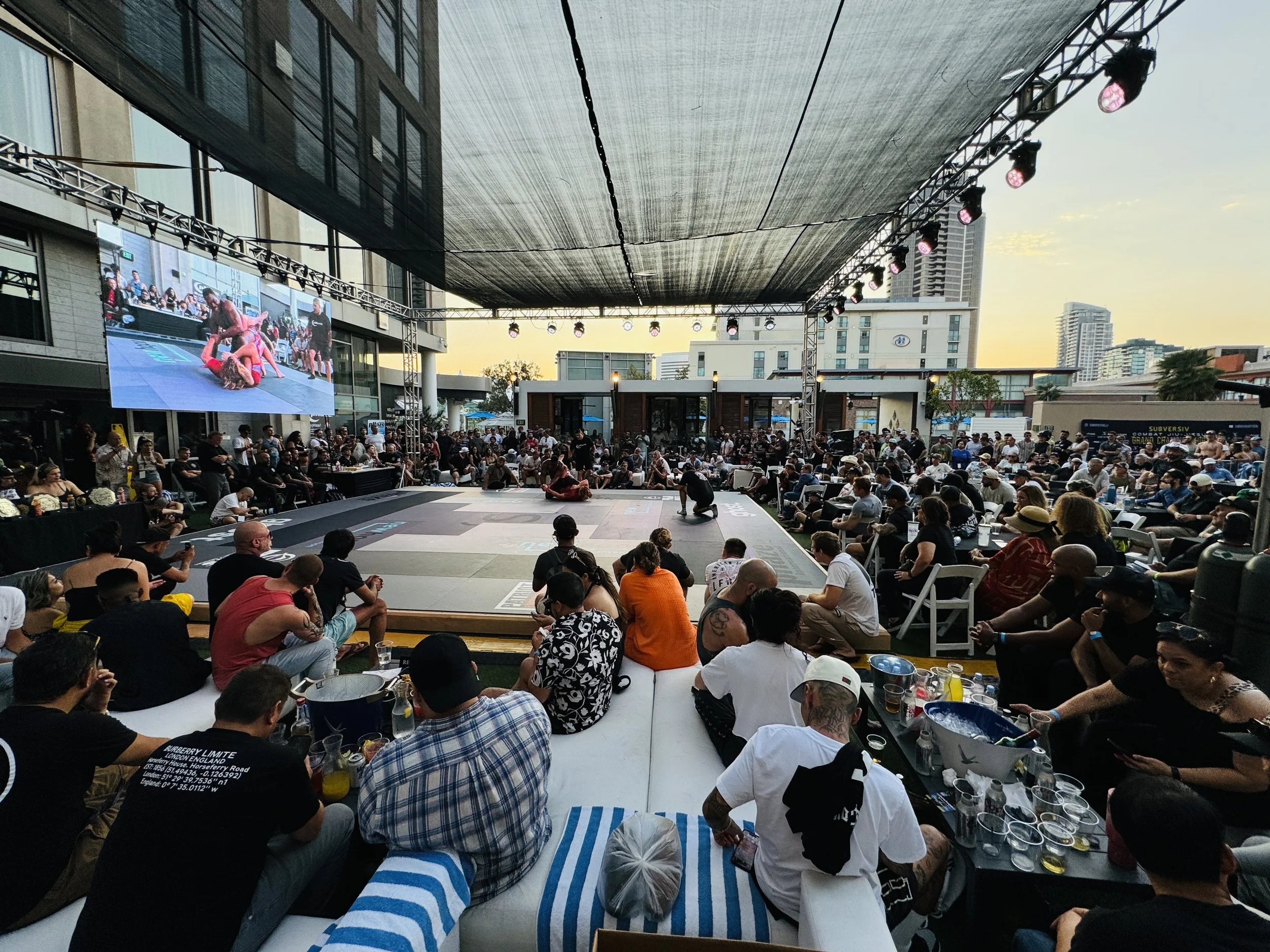 Large outdoor event venue with a stage and many attendees. There is a big screen showing martial arts fighters, and people are sitting at tables and standing around watching the event. The venue is decorated with lights and surrounded by tall buildings.