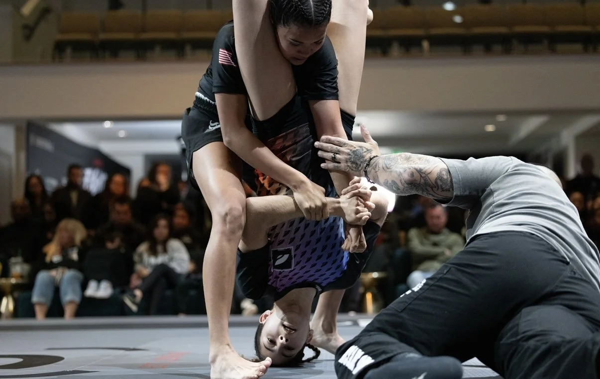 Two women engage in a Brazilian Jiu-Jitsu match where one woman is executing a submission move on the other, with a crowd watching in the background.