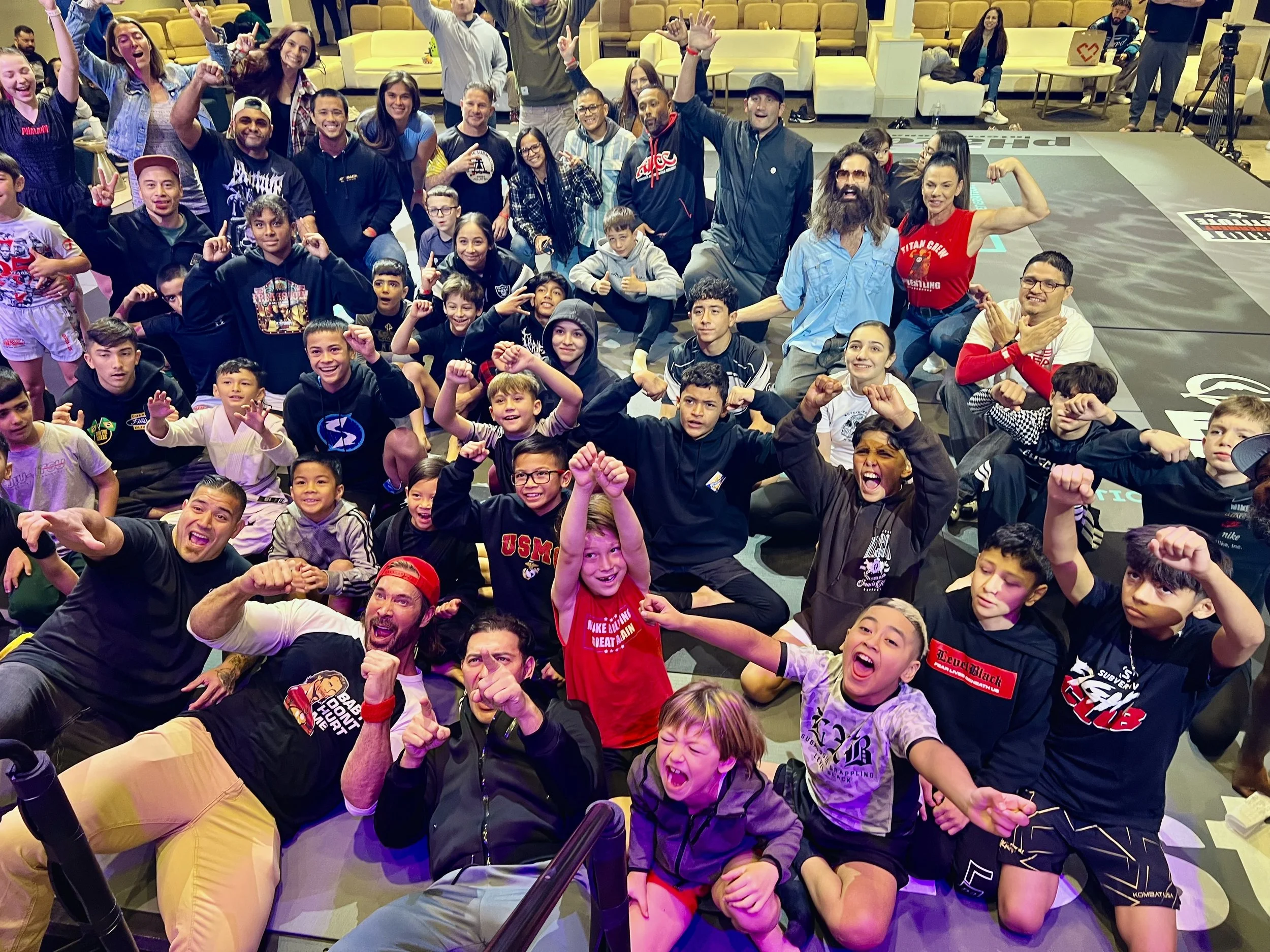 A large group of diverse people, including children and adults, gathered together indoors, smiling, many with their fists raised or making victory signs. They appear excited and happy, possibly celebrating or participating in a sporting or community event.