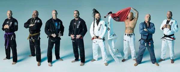 A group of nine men in various martial arts and Brazilian Jiu-Jitsu uniforms standing against a plain background.