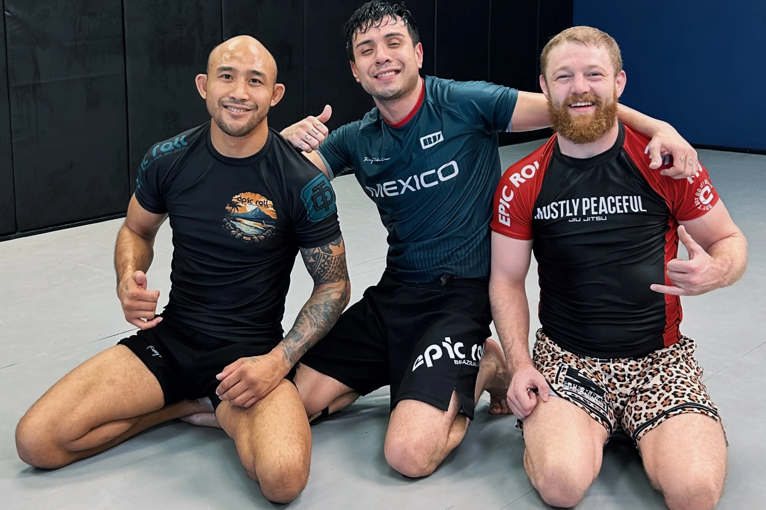 Three men sitting on the floor of a martial arts gym, smiling and posing for the camera, with two of them giving thumbs up. They are dressed in athletic shirts and shorts, with tattoos and a beard visible on one of them.