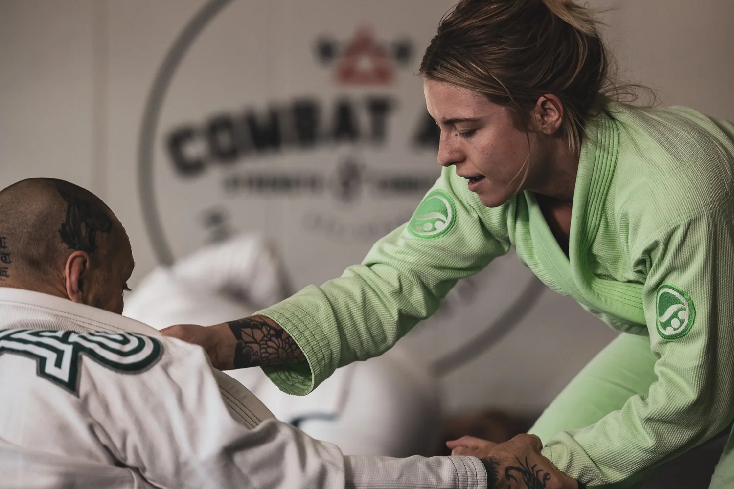 A woman in a neon green jiu-jitsu gi helping an injured man in a white gi, in a gym with a logo that says 'GENTAL' behind them.